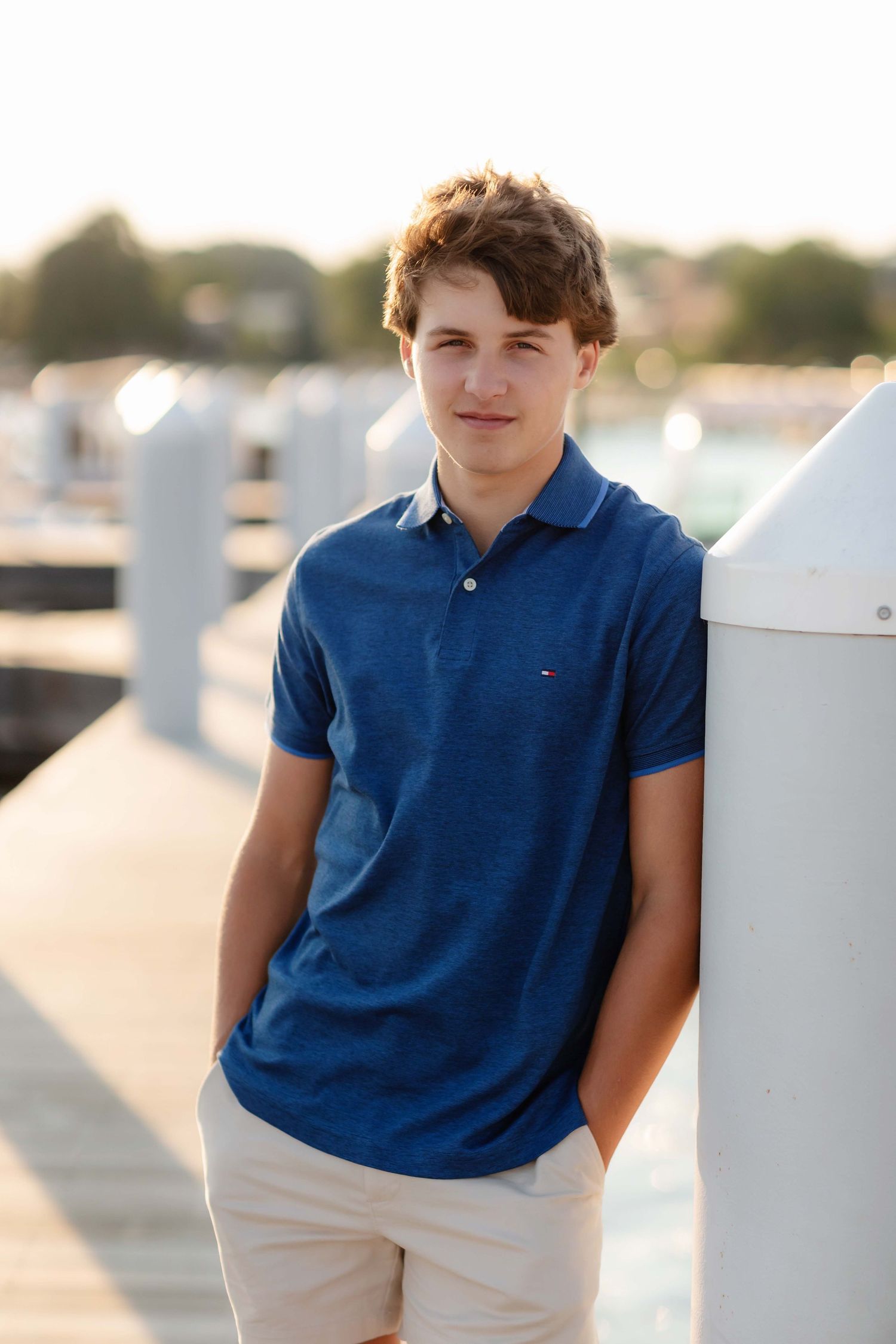 Teen boy in blue Tommy Hilfiger polo leaning against white pier post at a sunny marina dock.