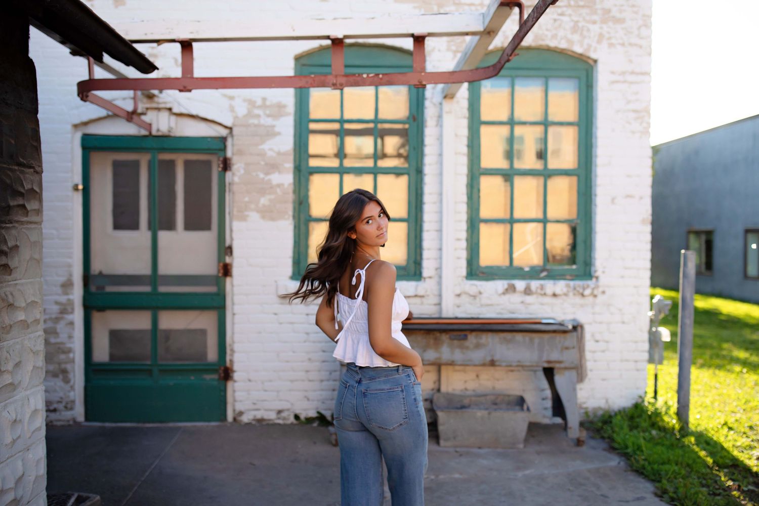 Young woman in white top and jeans posing outside a rustic white brick building with green-trimmed windows.
