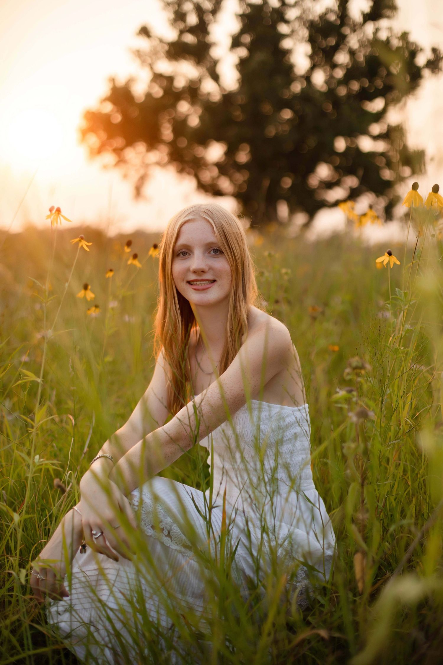 Young woman in white dress sitting in wildflower meadow at golden sunset with warm light and yellow flowers.