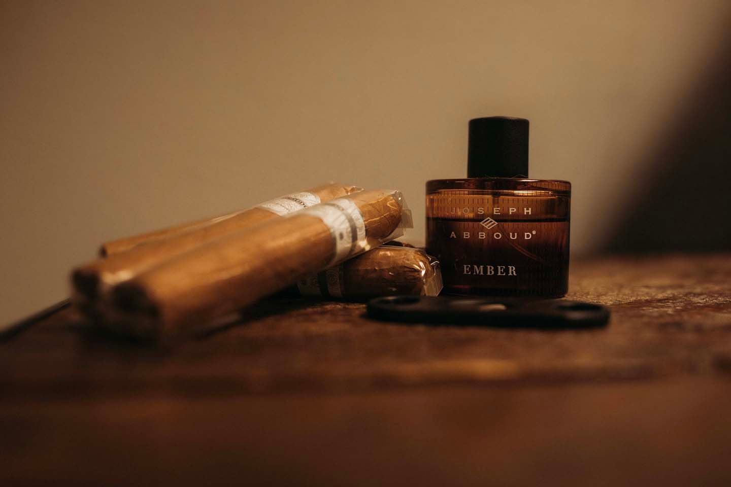 Ember cologne bottle beside cigars and black scissors on a rustic wooden surface in warm moody lighting.