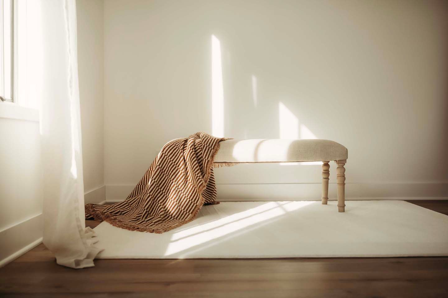 A minimalist white bench with wooden legs, draped with a cozy knit blanket, sits on a cream rug in a sunlit room.