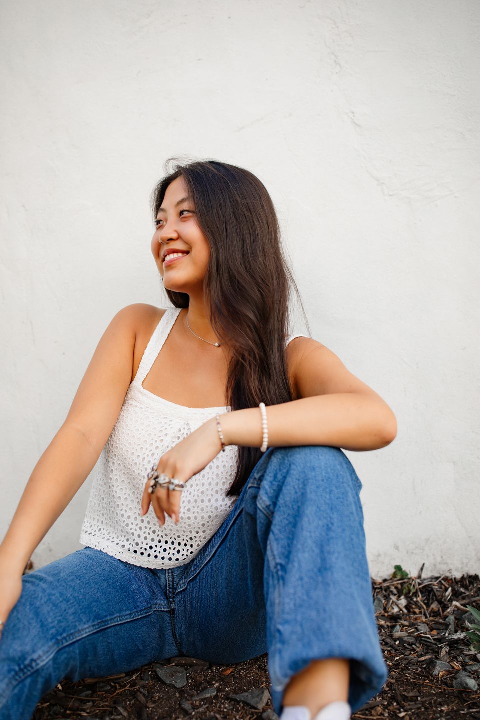 Young woman in white eyelet top and jeans sits casually against white wall, looking to the side with a bright smile.