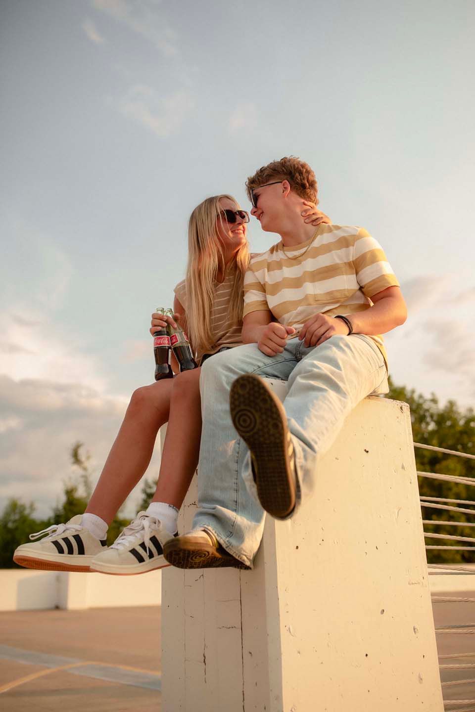 Young couple sitting on a concrete ledge outdoors, sharing drinks and smiling at each other in warm sunset light.