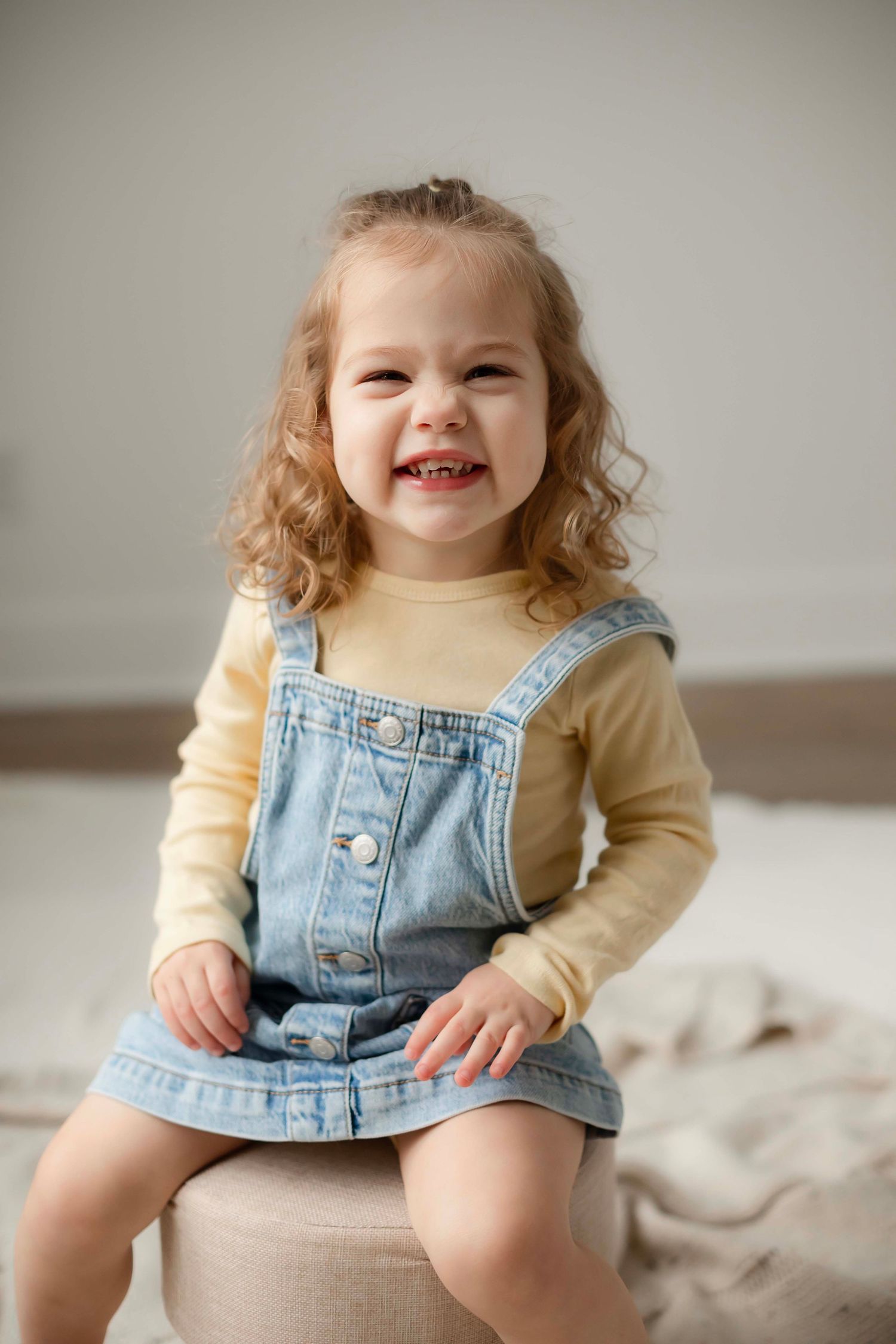 A child in denim overalls and yellow sweater sits on a light colored couch in a progression of candid poses.