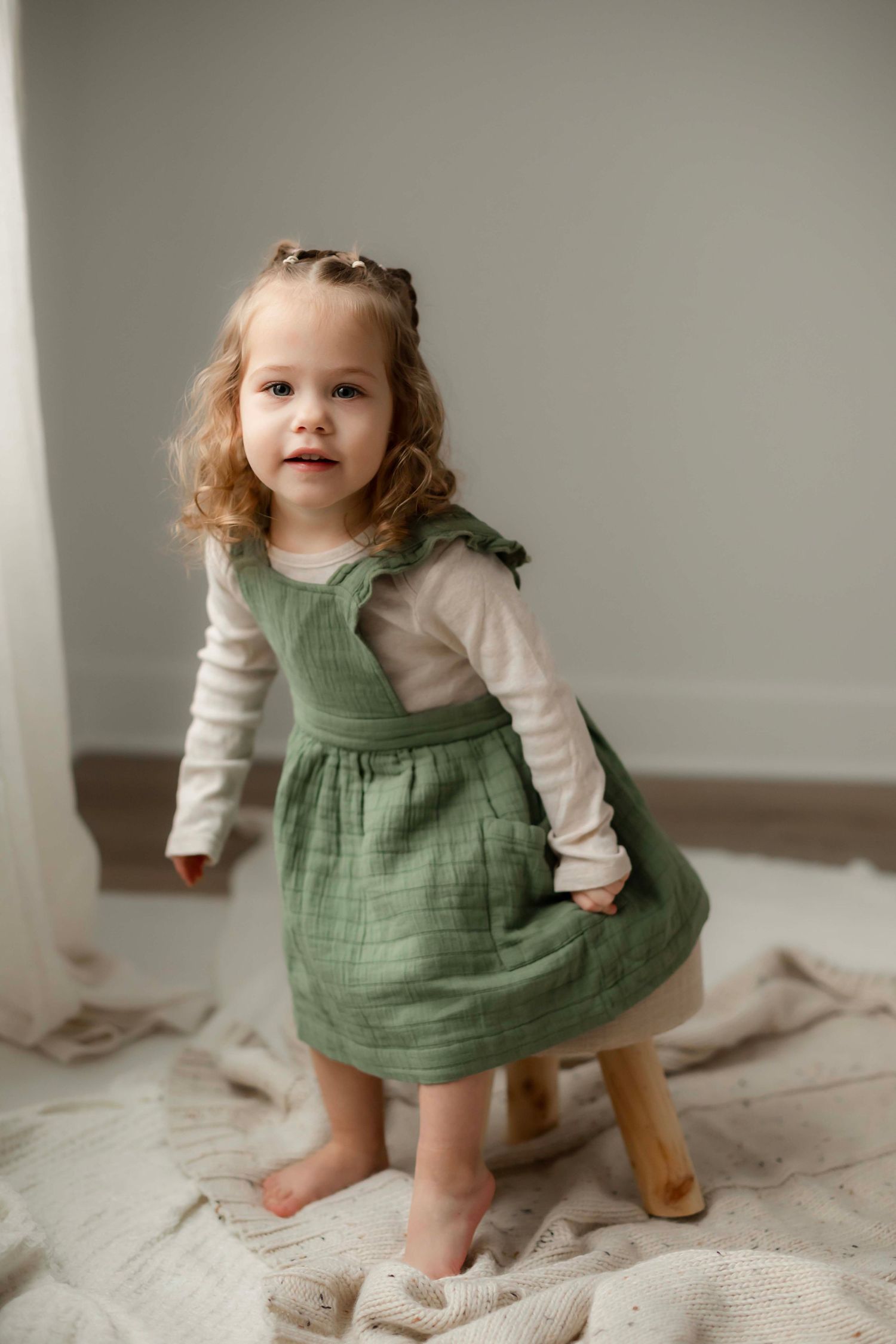 Small child wearing a sage green pinafore dress and cream long sleeve shirt stands on white textured bedding.