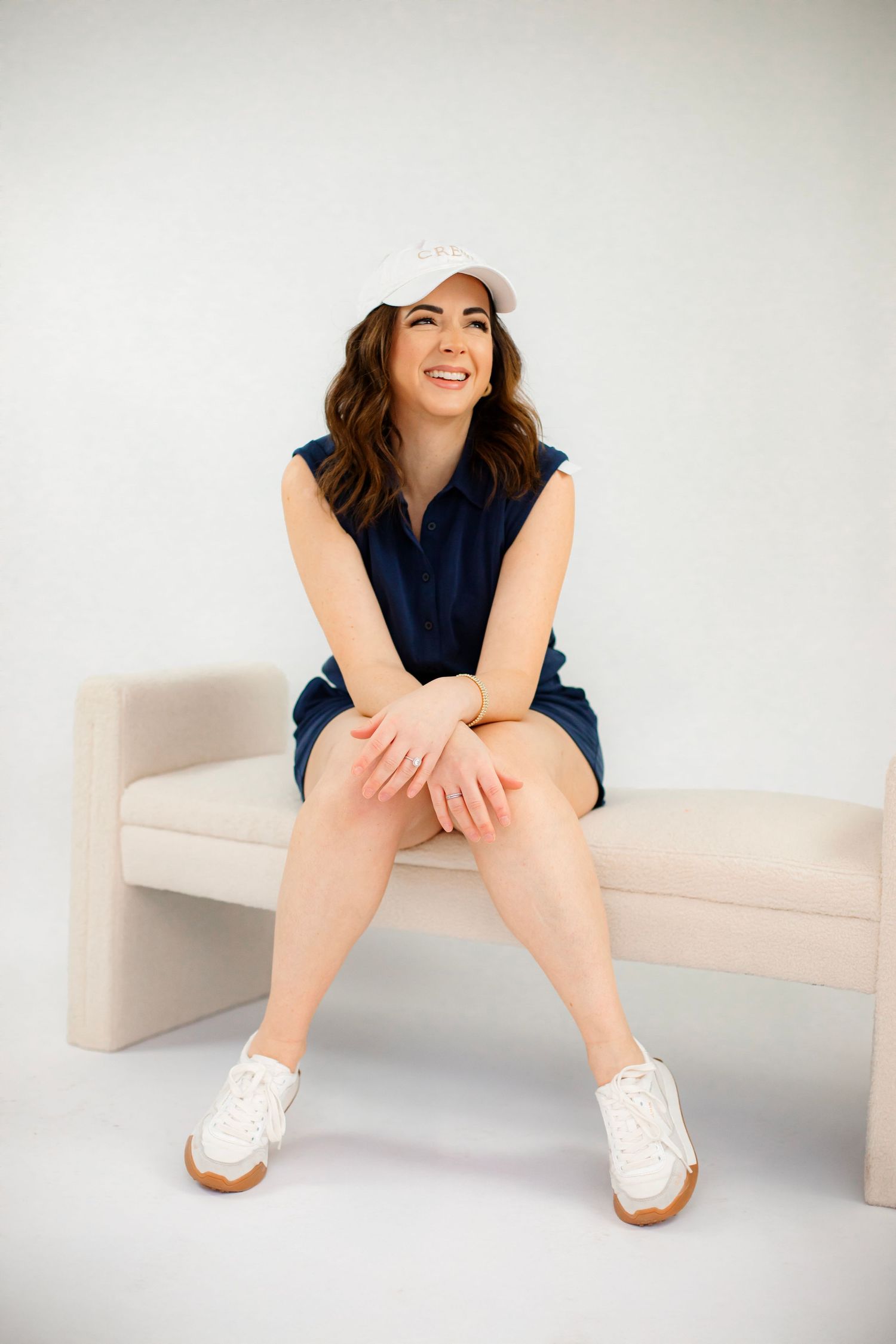 Woman in navy romper and baseball cap sits casually on white bench.