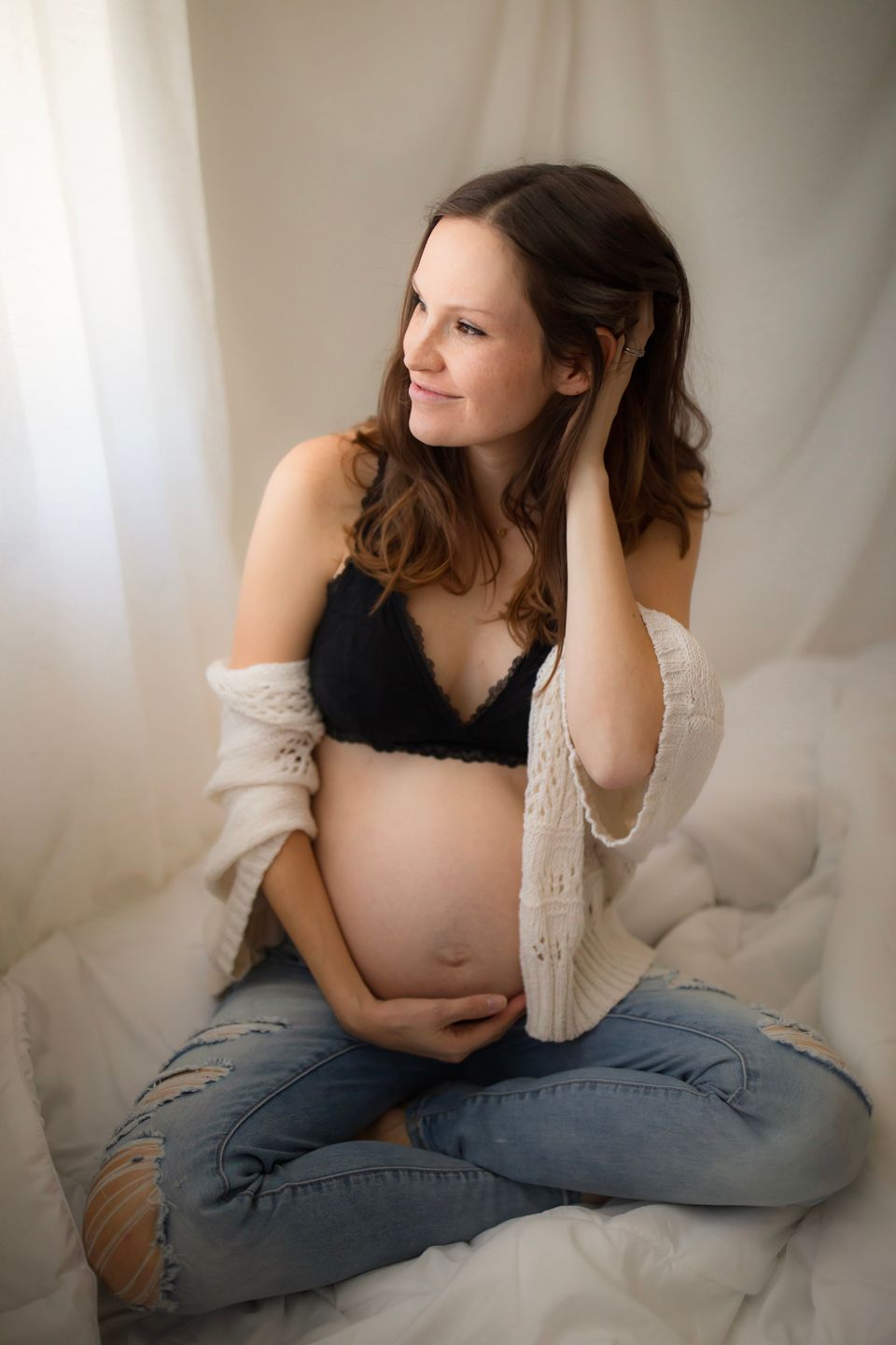 Expectant mother in jeans and black bralette poses tenderly with her baby bump against white backdrop.