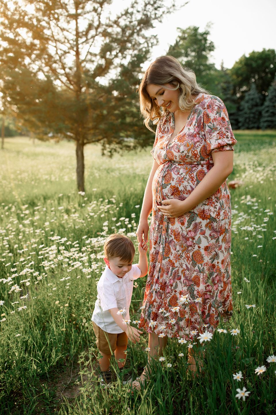 A floral dress-clad woman and small child stand together in a sunlit meadow filled with white wildflowers at sunset.
