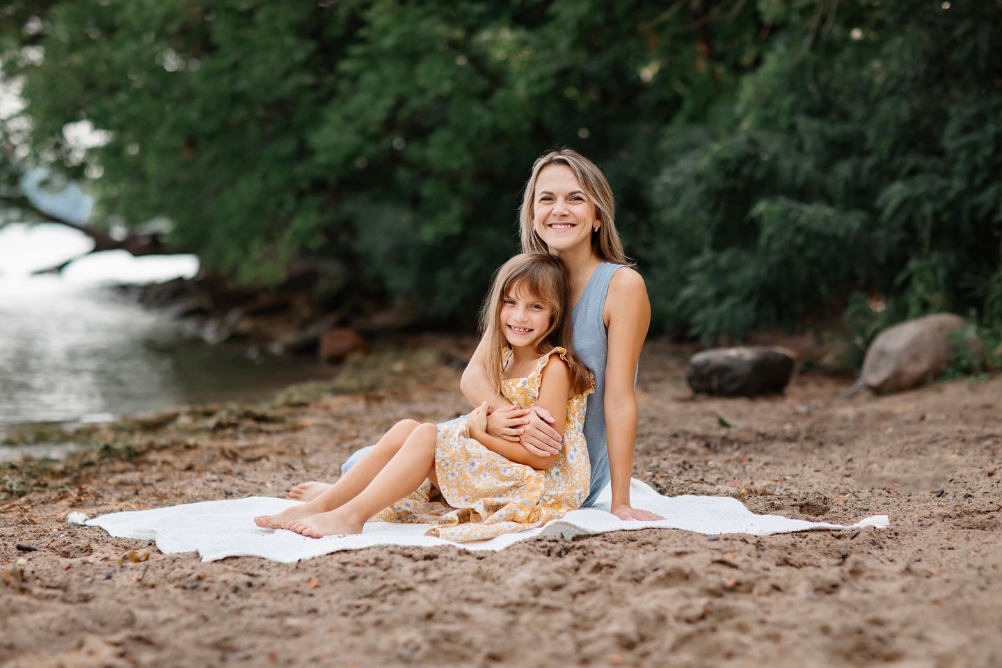 A parent and child sit together on a white blanket by the rocky riverbank during a summer outdoor portrait session.