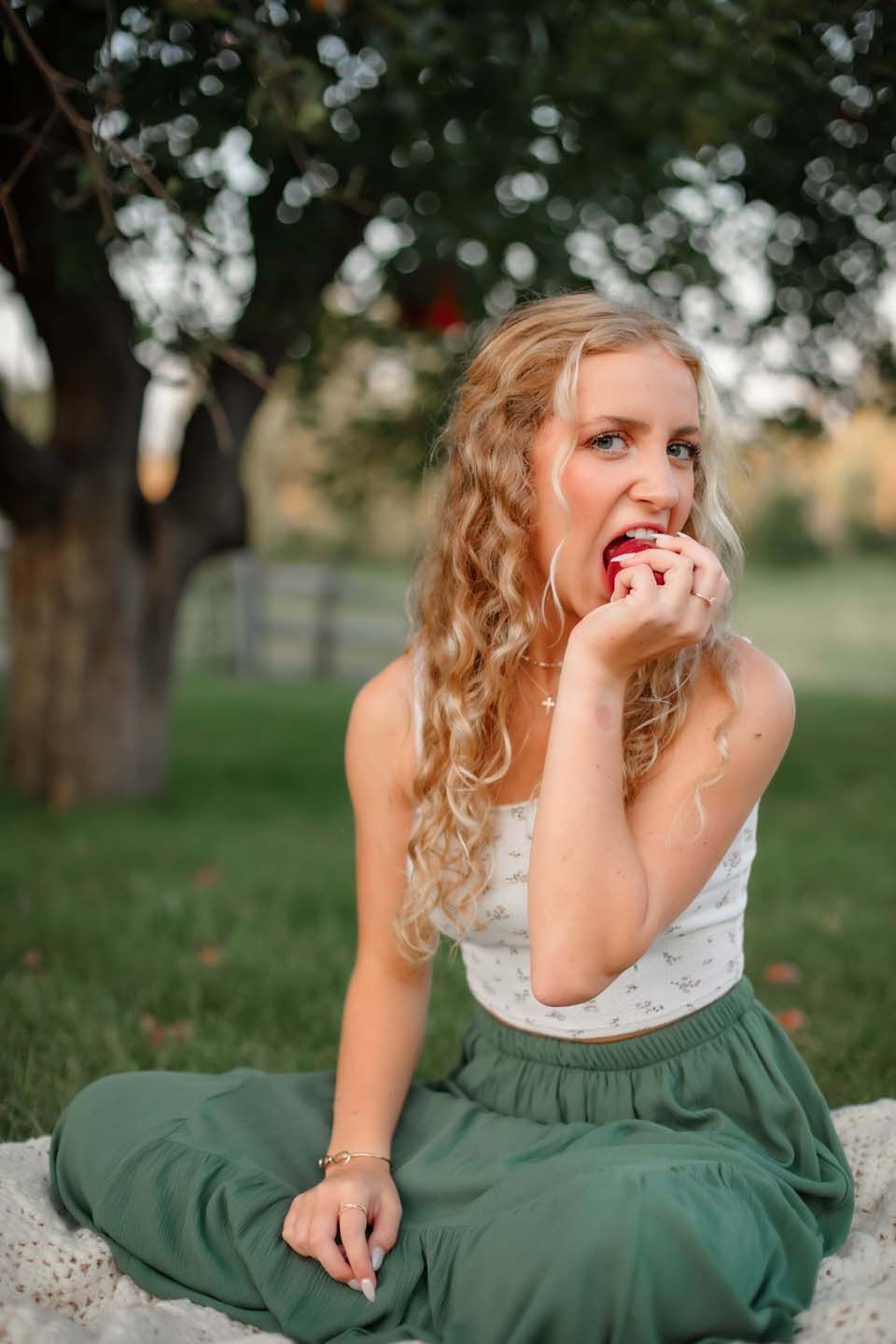 Portrait in green skirt and white top sitting on blanket under bokeh tree lighting.