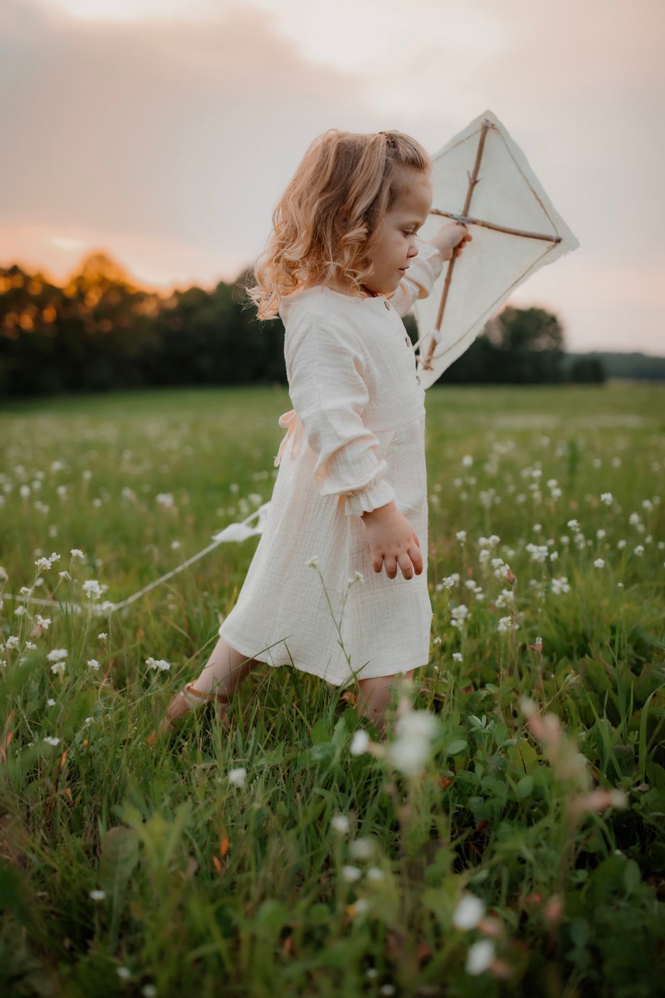 Child in white dress holding a handmade kite in a wildflower meadow at sunset.