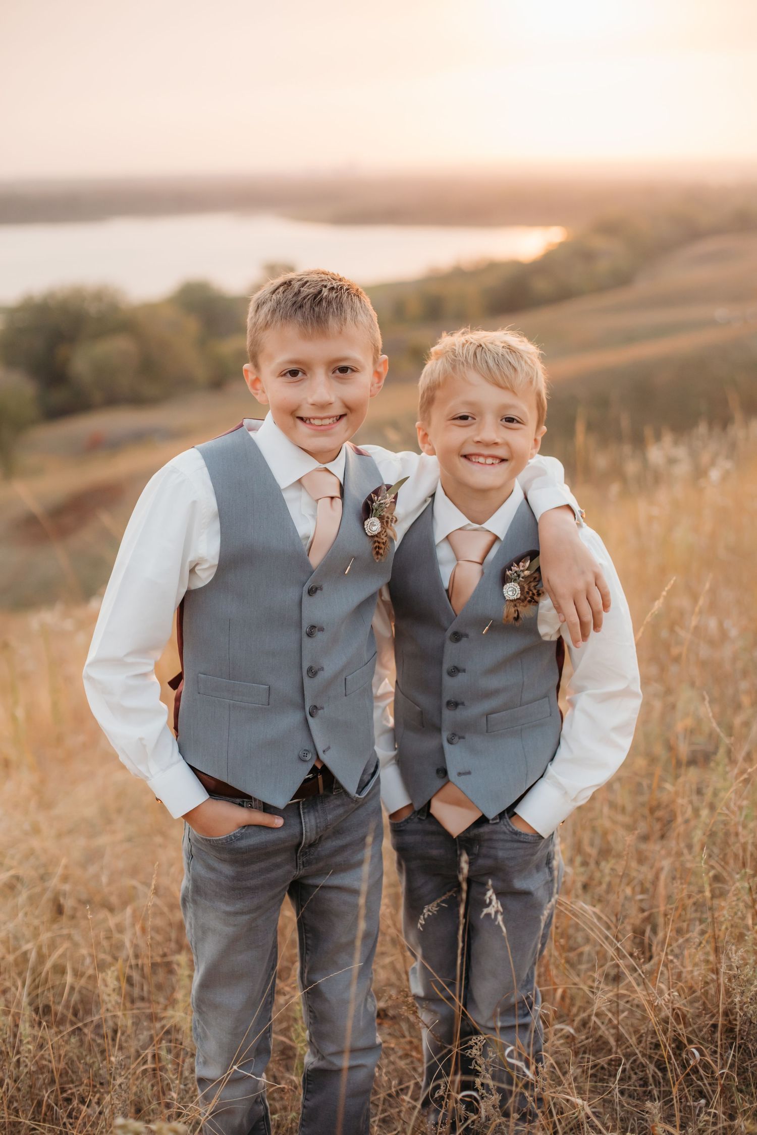Two young boys in matching grey vests pose together on grassy hillside at sunset.