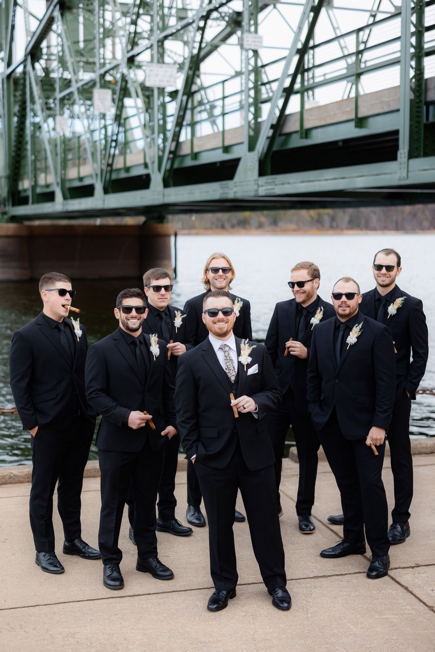 Groomsmen in black suits and sunglasses pose confidently under a bridge for wedding photos.