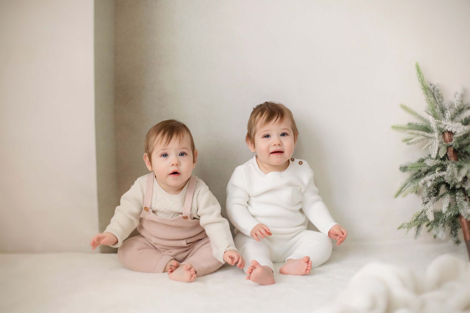 Two babies in coordinating outfits sit together on white surface with winter decor.