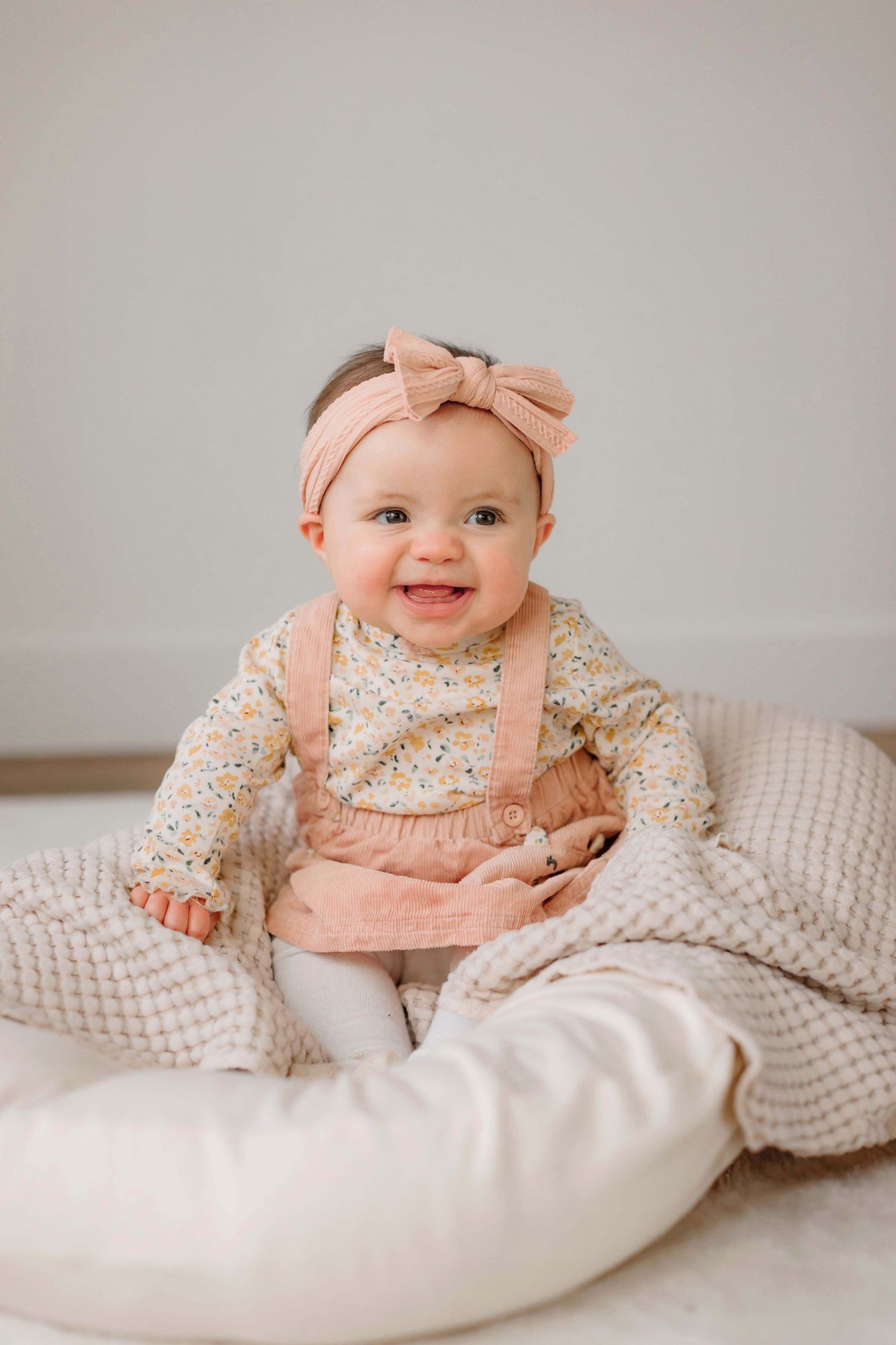 Baby in pink overalls and floral top sitting on white cushions.