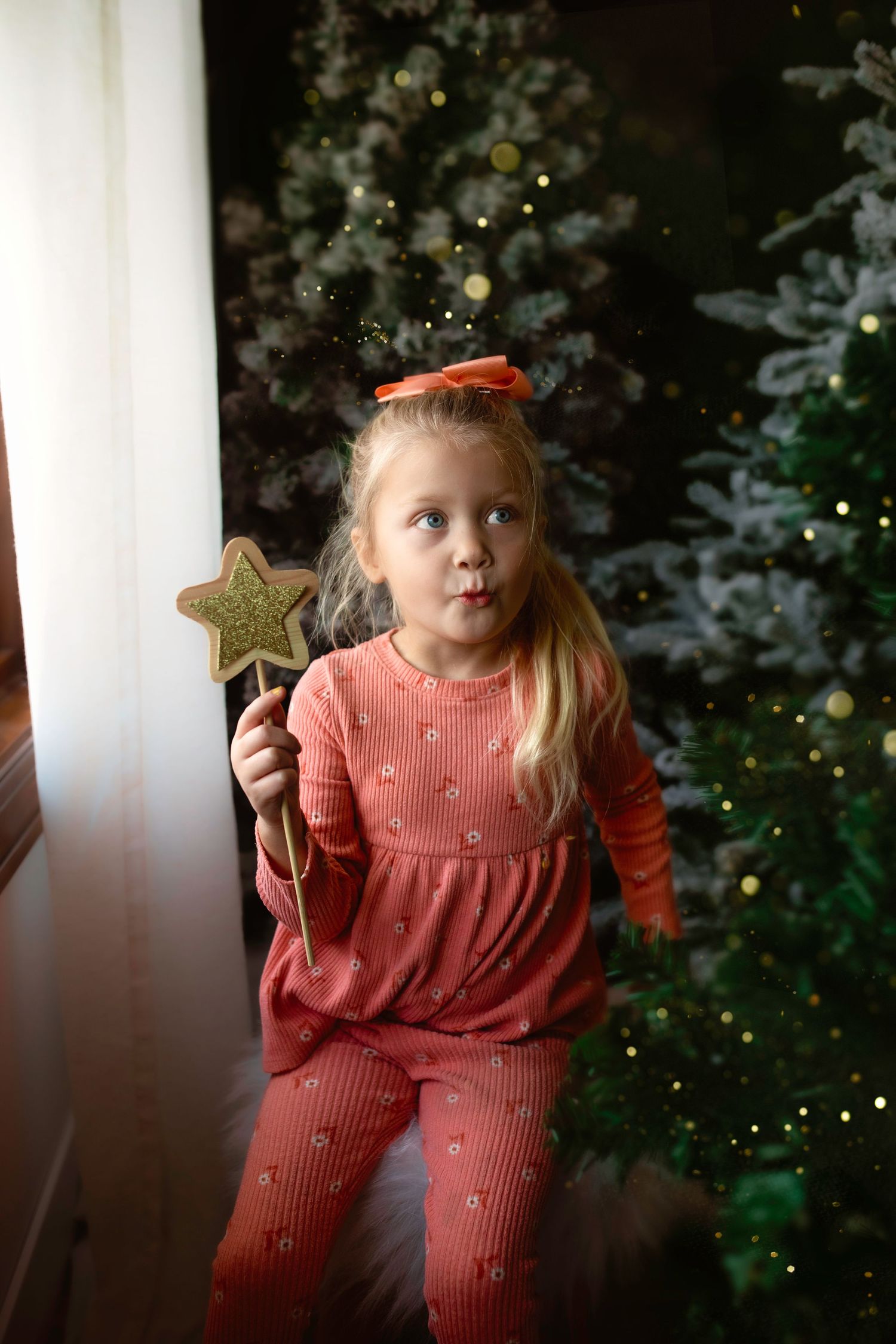Child in coral outfit holding gold star wand in front of decorated Christmas tree.