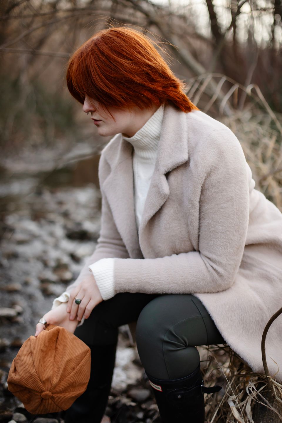 A figure in a beige coat and white turtleneck sits pensively outdoors with vibrant red hair against an autumn backdrop.