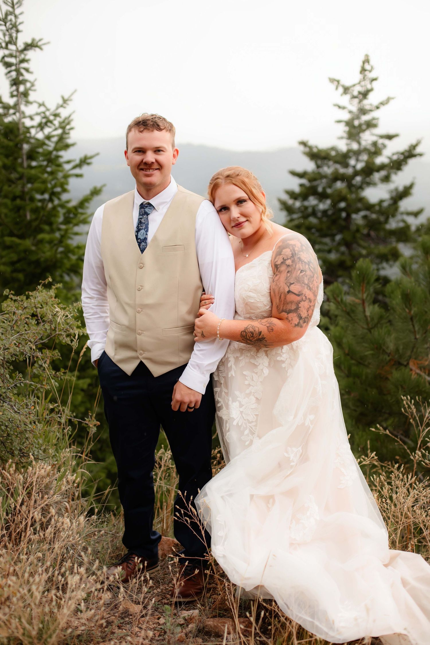A couple poses on a mountain trail wearing wedding attire with evergreen trees and mountains in the background.