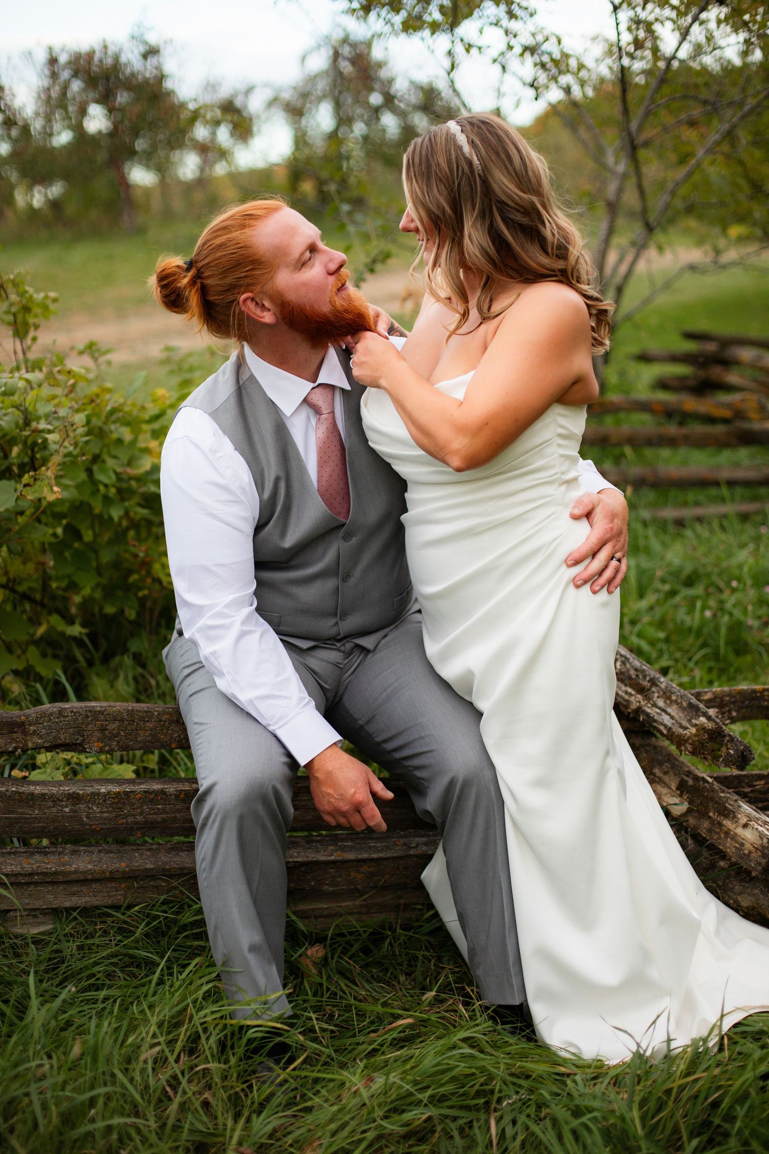 A couple shares an intimate moment outdoors on their wedding day, with a rustic wooden fence and greenery in the background.