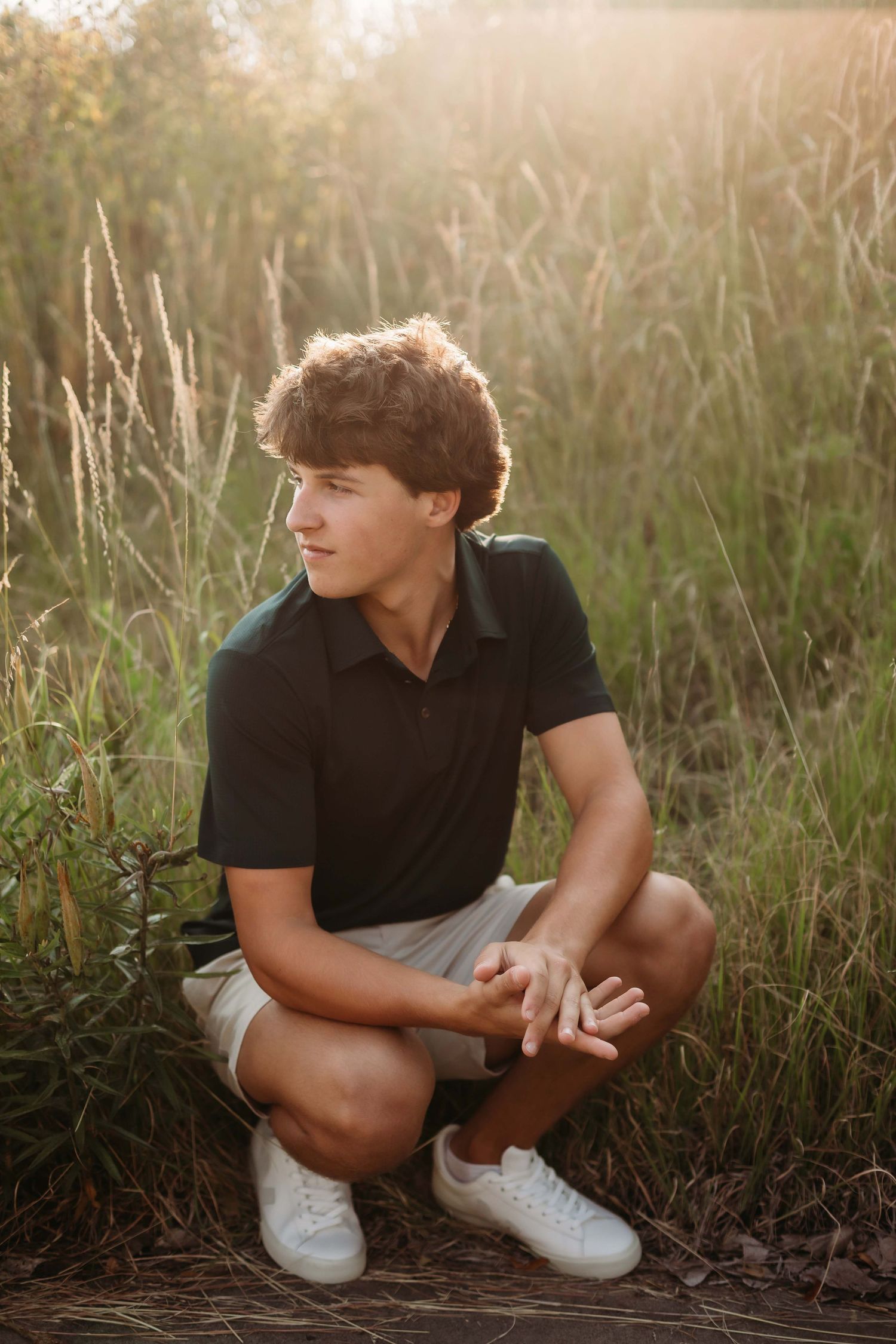 Young man with curly hair crouching in a sunlit field, wearing a black polo shirt and khaki shorts.