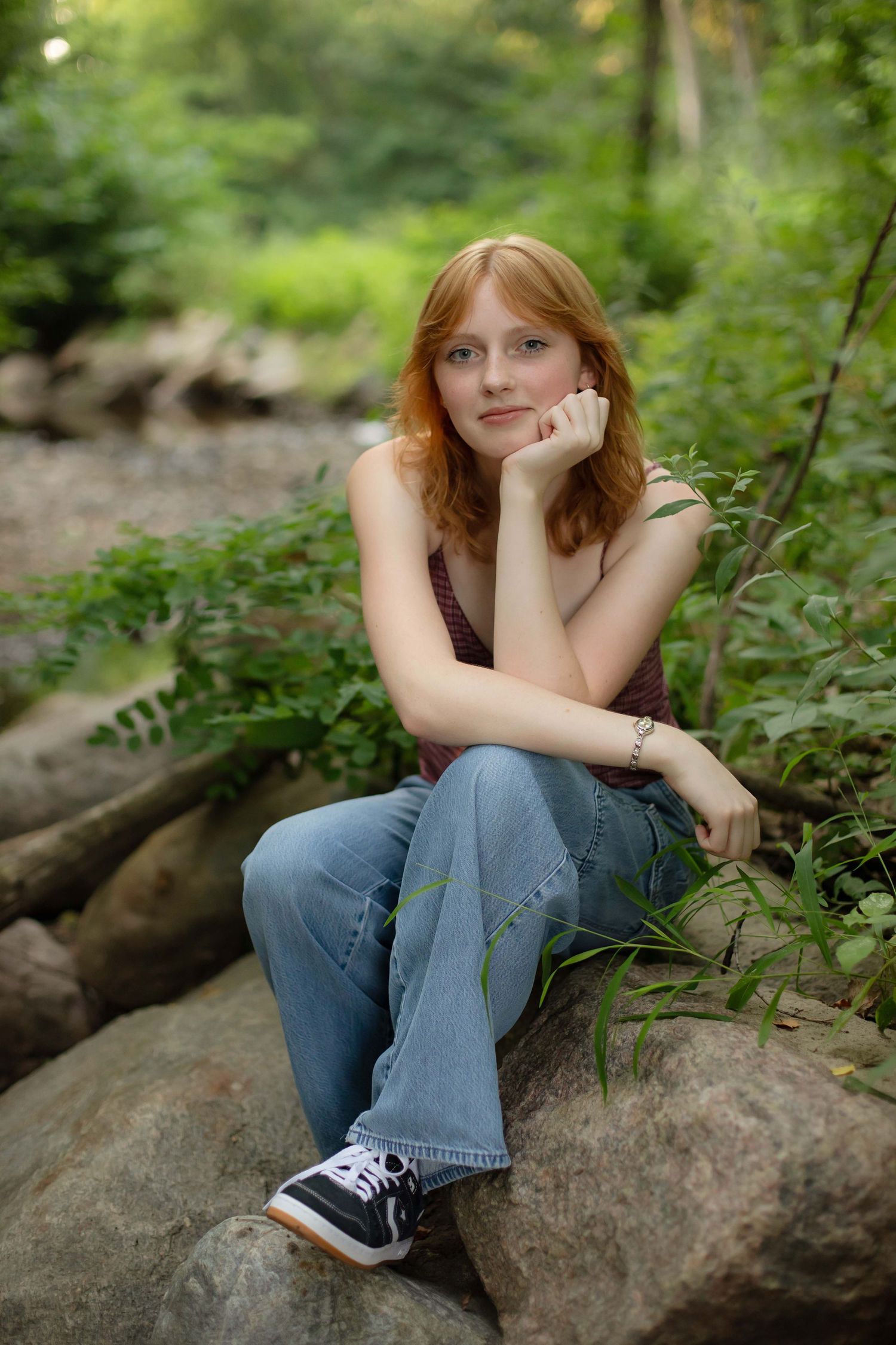 Teenage girl with red hair sitting on rocks in a lush green forest, resting chin on hand in a thoughtful pose.