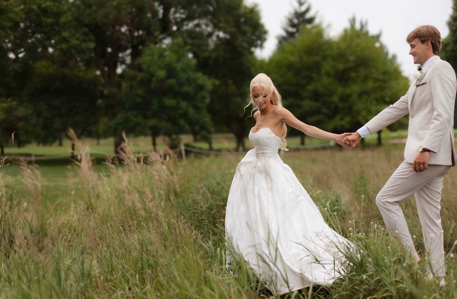 Bride and groom holding hands in a lush green meadow, bride in white gown, groom in light suit.