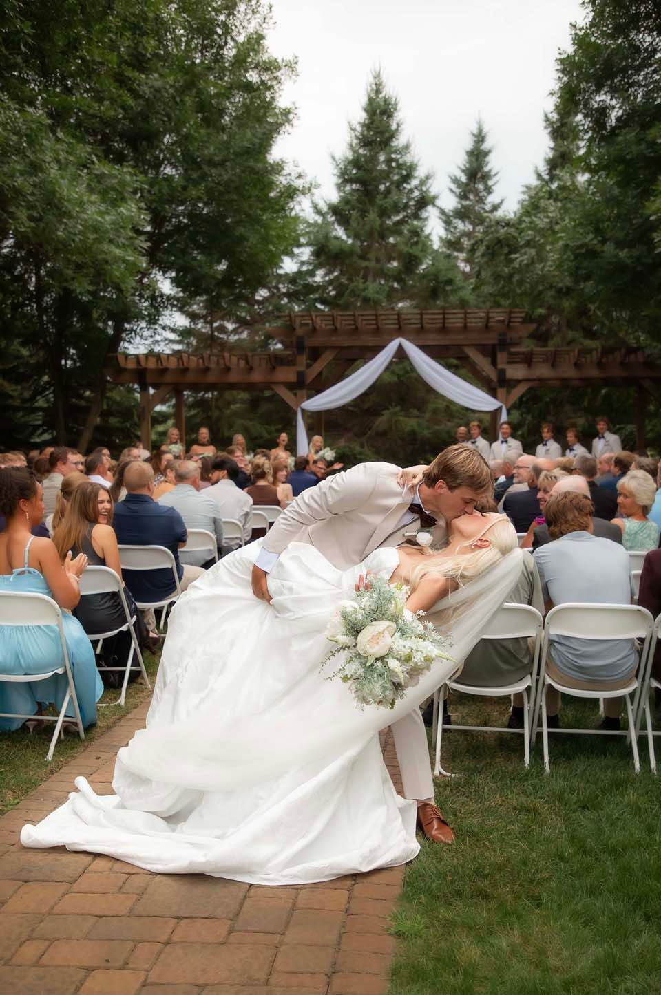 Groom dips bride for a kiss at outdoor wedding ceremony with seated guests and wooden arbor backdrop.