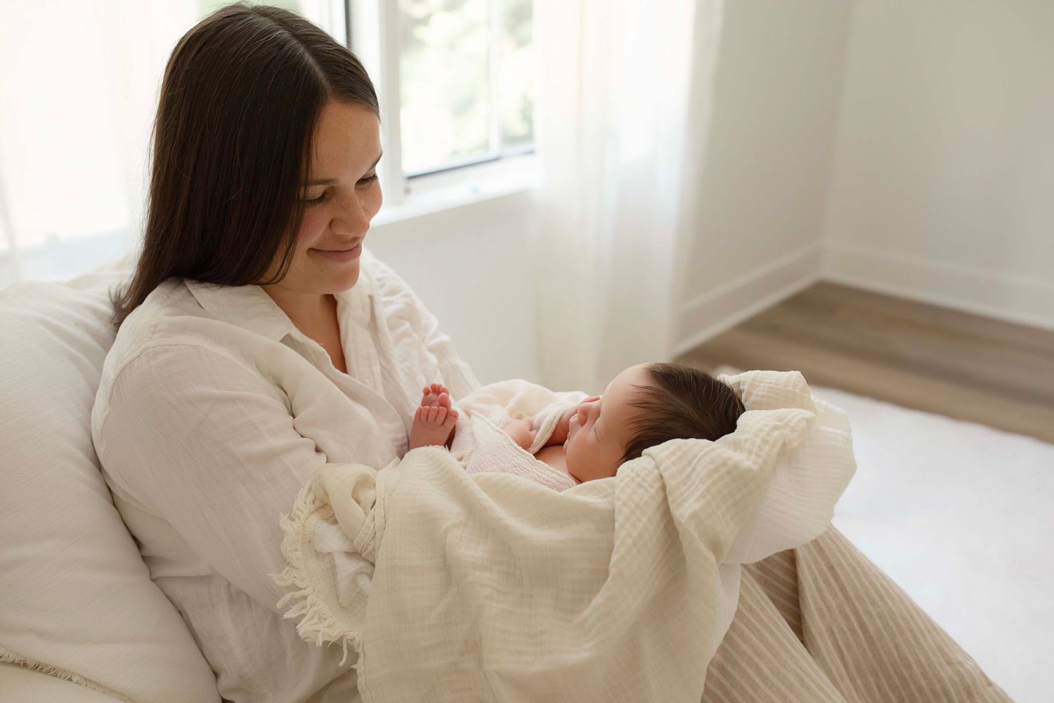 A mother in a white robe tenderly holds her sleeping newborn baby wrapped in a soft white blanket indoors.