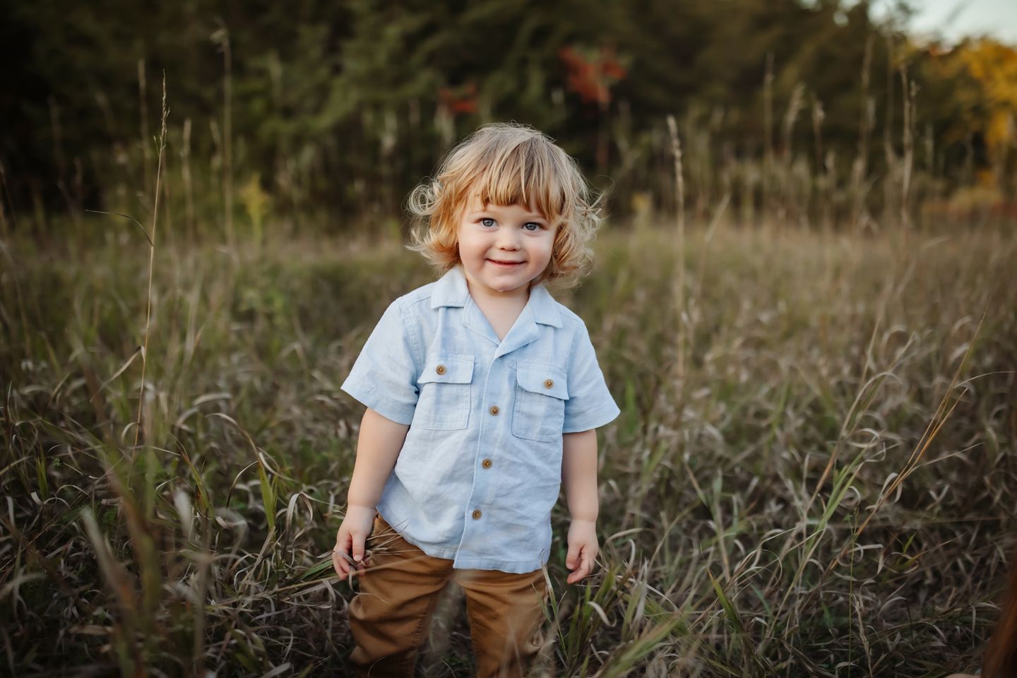 Young child in light blue buttoned shirt and brown pants standing in tall grass during golden hour in autumn field.
