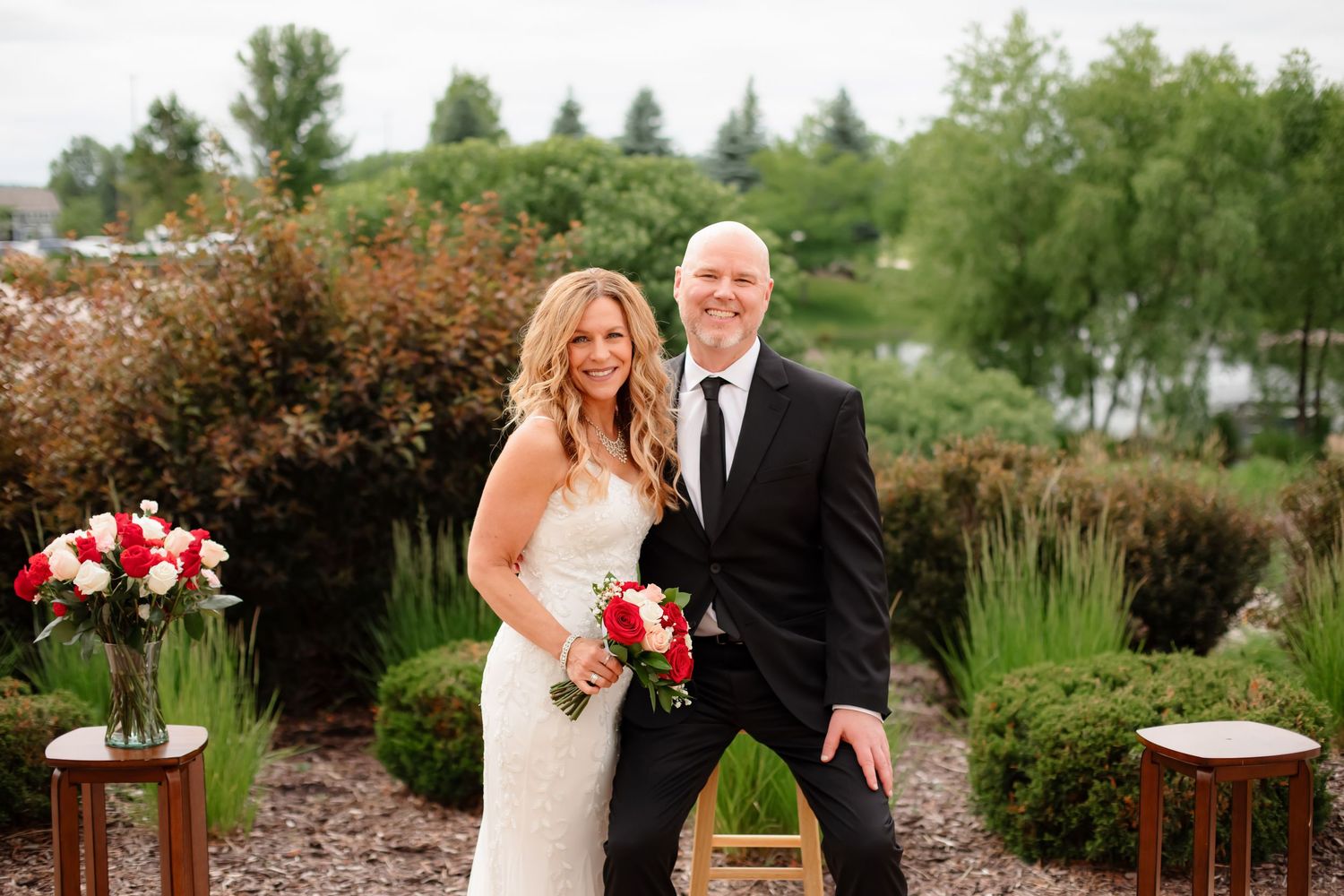 Wedding party members gather near a decorative gazebo with greenery and flowers in the background.