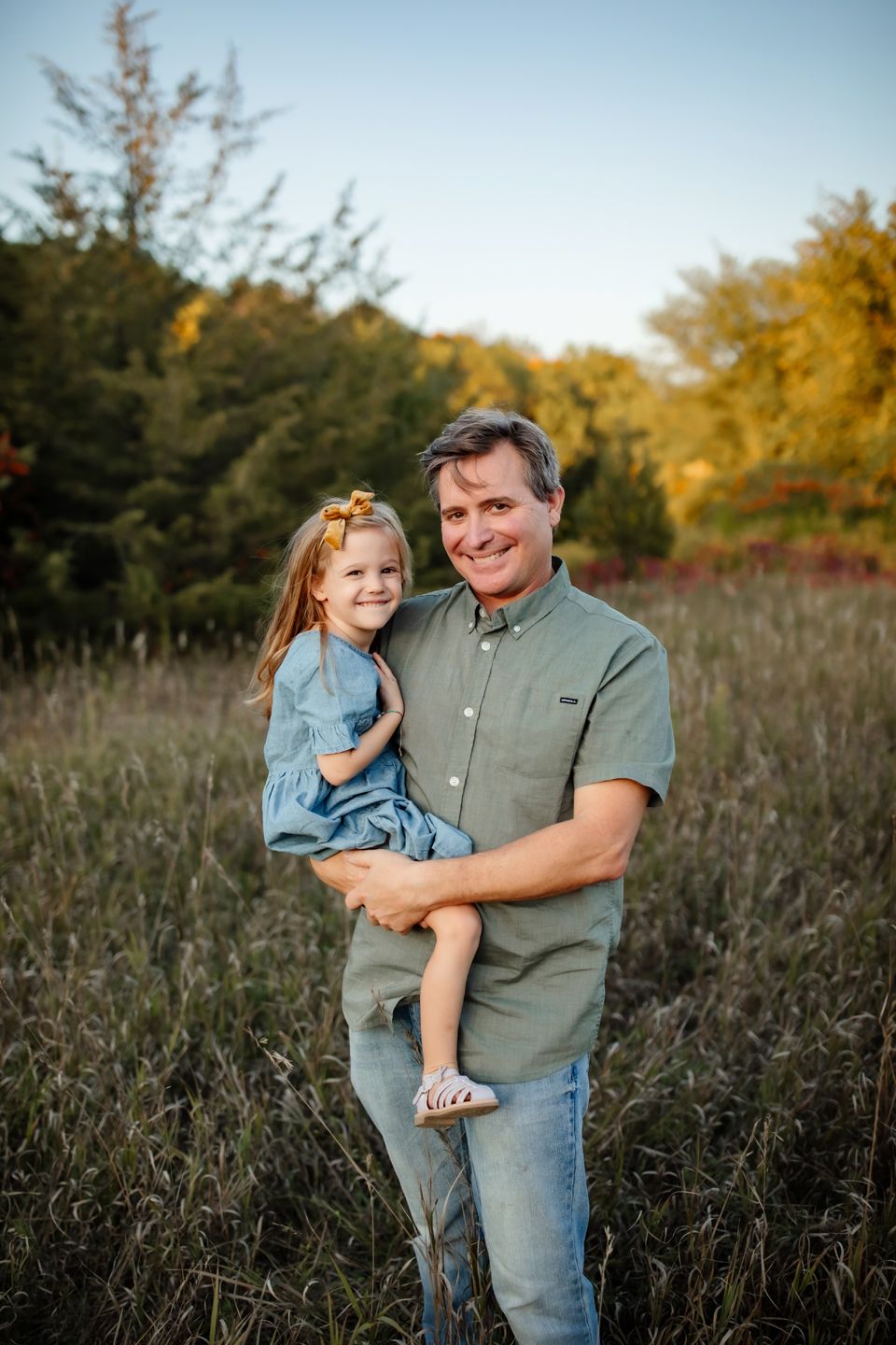 Person in casual attire holding a small child in a field during golden hour.
