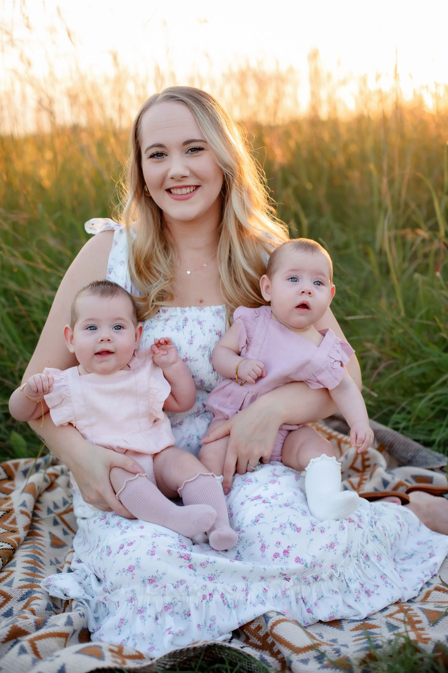 Mother sitting with twin babies on blanket in sun-drenched field during golden hour.