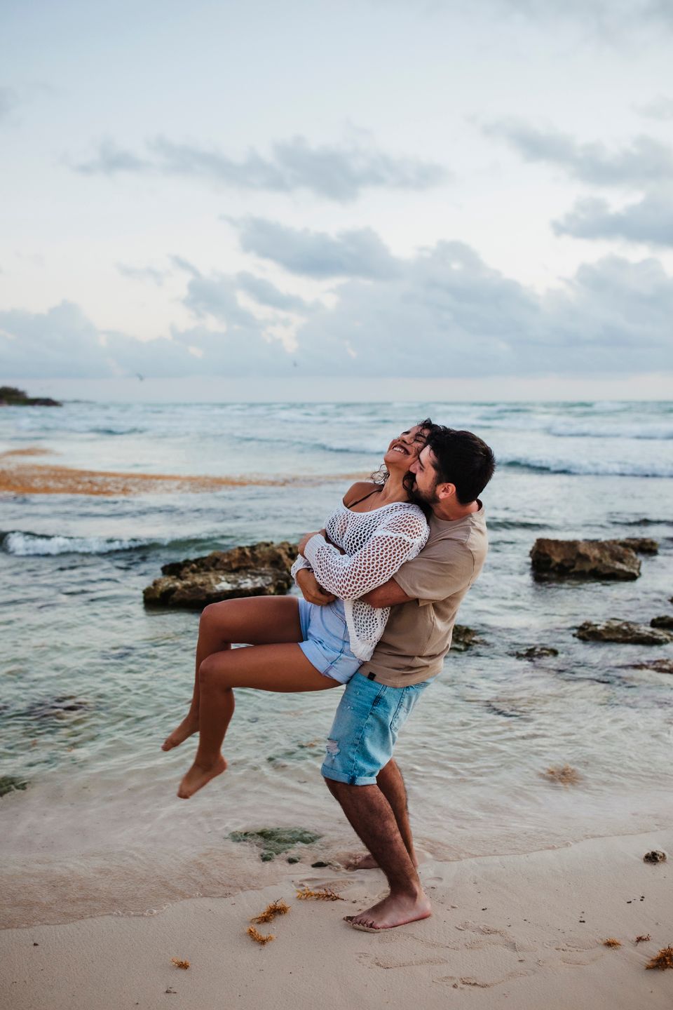A playful beach moment as one person lifts the other in front of crashing waves and rocky coastline.
