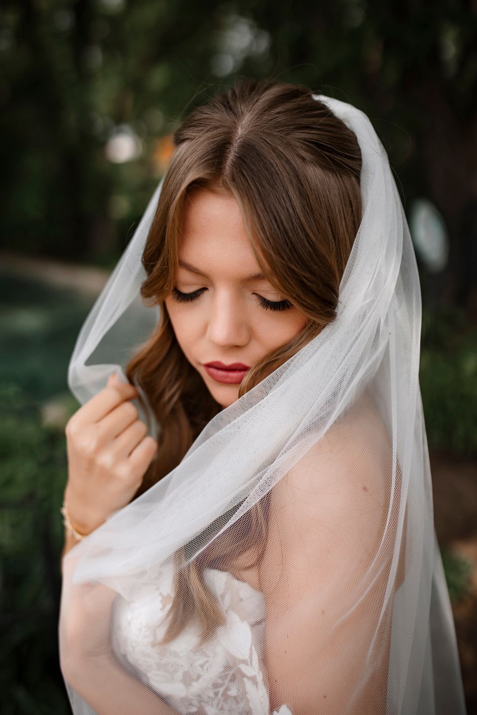 Close-up portrait of bride with flowing veil and red lipstick against blurred natural background.