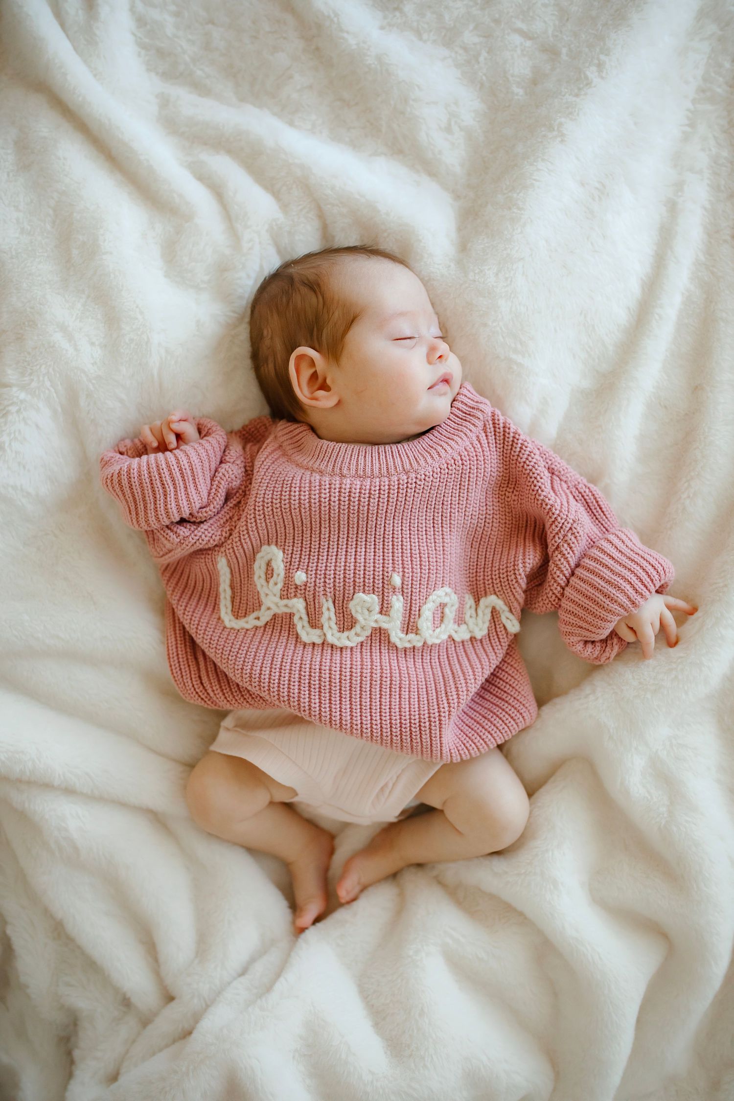 Sleeping baby wearing pink knitted sweater with name embroidered lying on white blanket.