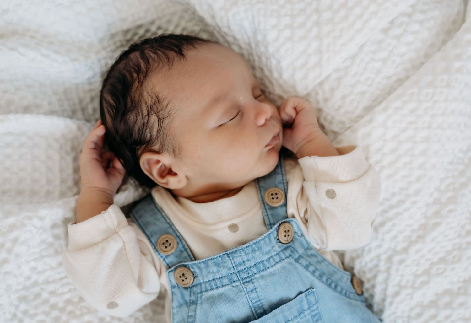 Peaceful sleeping baby in blue denim overalls and cream colored top on white bedding.