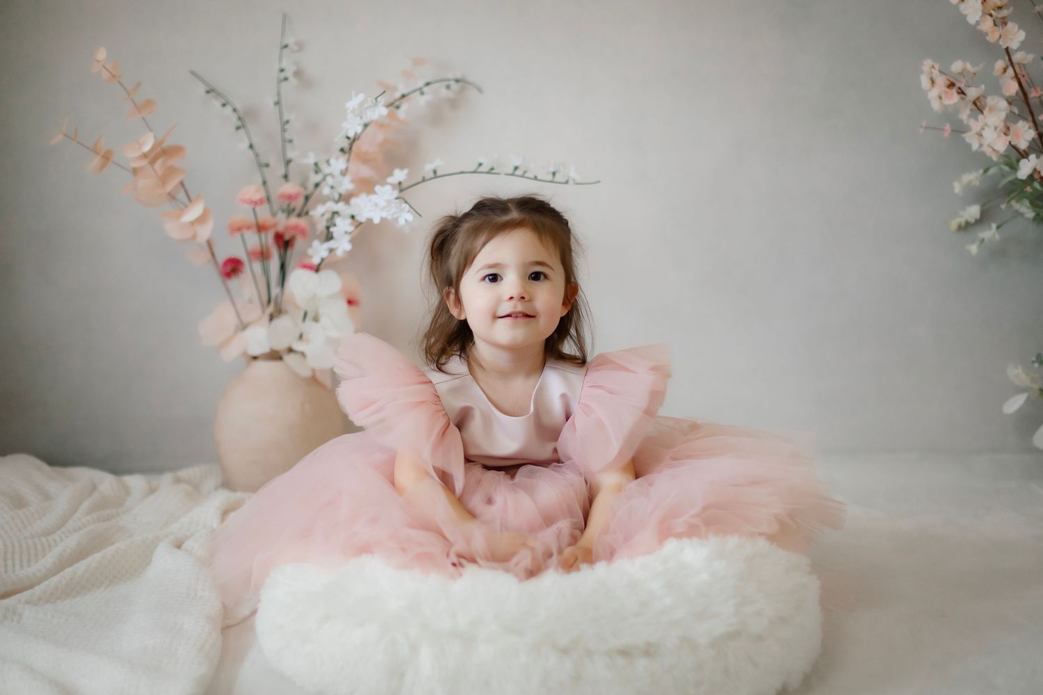 Child in pink tulle dress poses on white cushion with floral decorations.