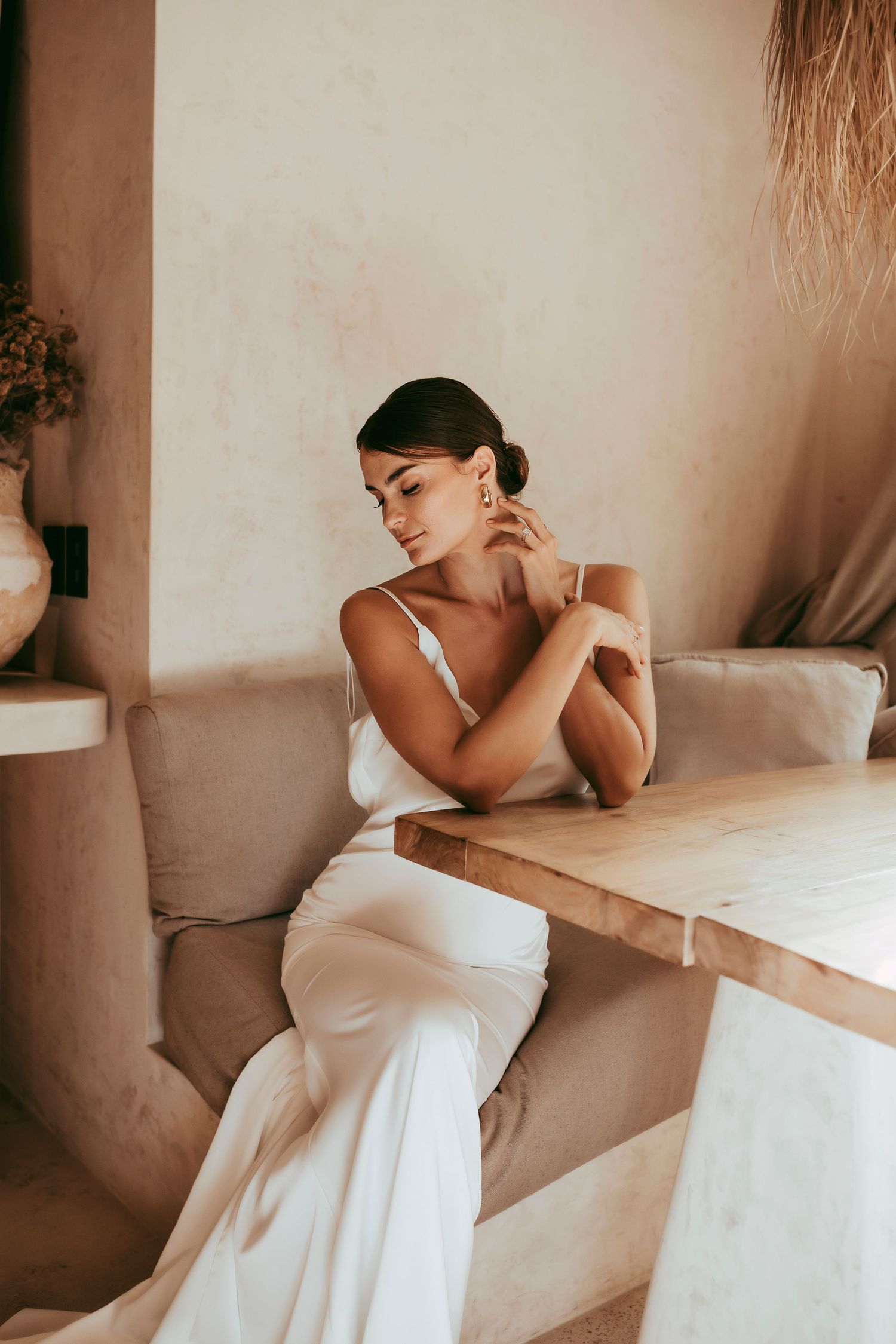 Woman in white slip dress seated at wooden table in minimalist setting.