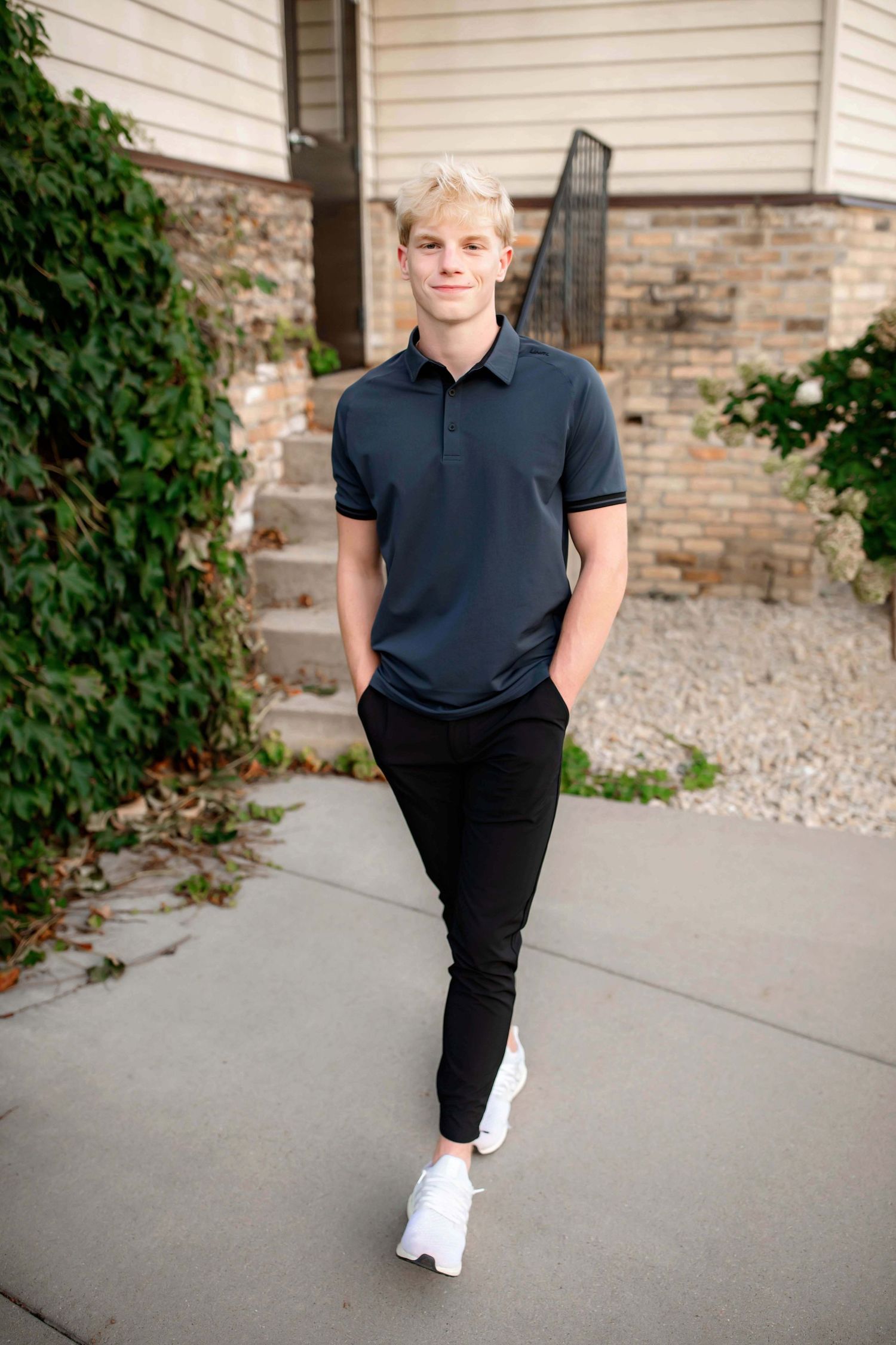 Casual outdoor portrait against white siding and brick wall wearing navy polo and white sneakers.