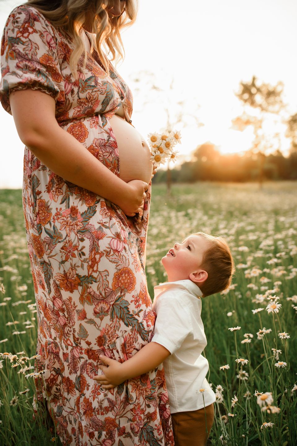 A pregnant woman in floral dress stands in a blooming meadow at sunset with a young child hugging her belly.