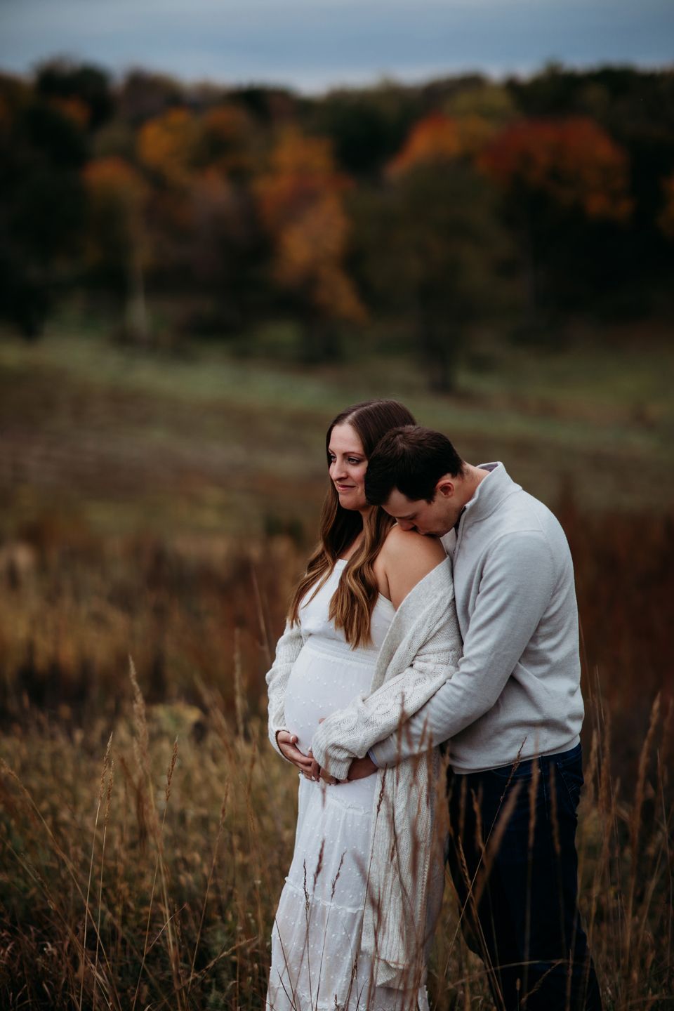 A couple shares an intimate moment in a field with autumn foliage in the background.
