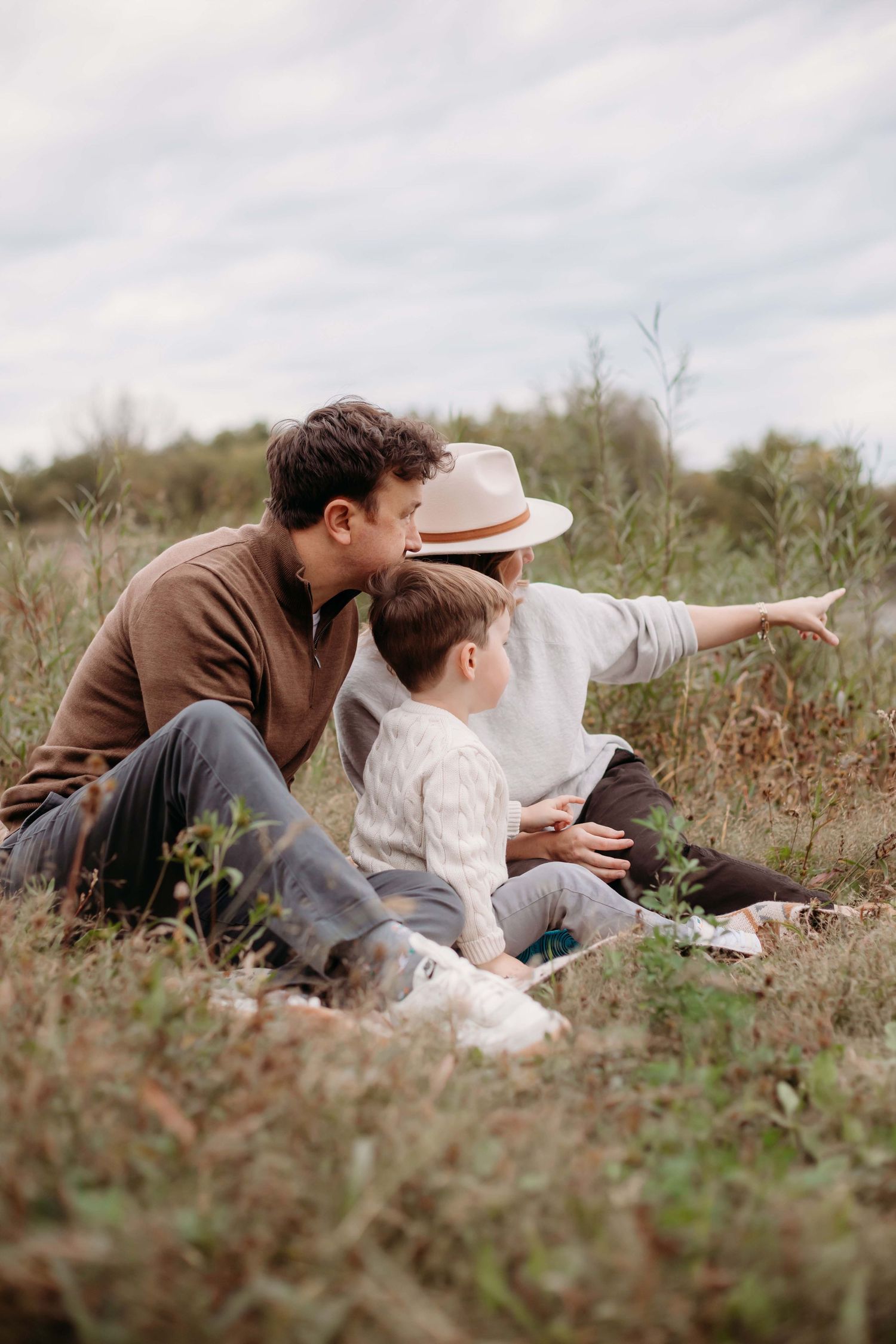 Family sitting in a field, with a person in a hat pointing at the horizon while a child and adult look on.