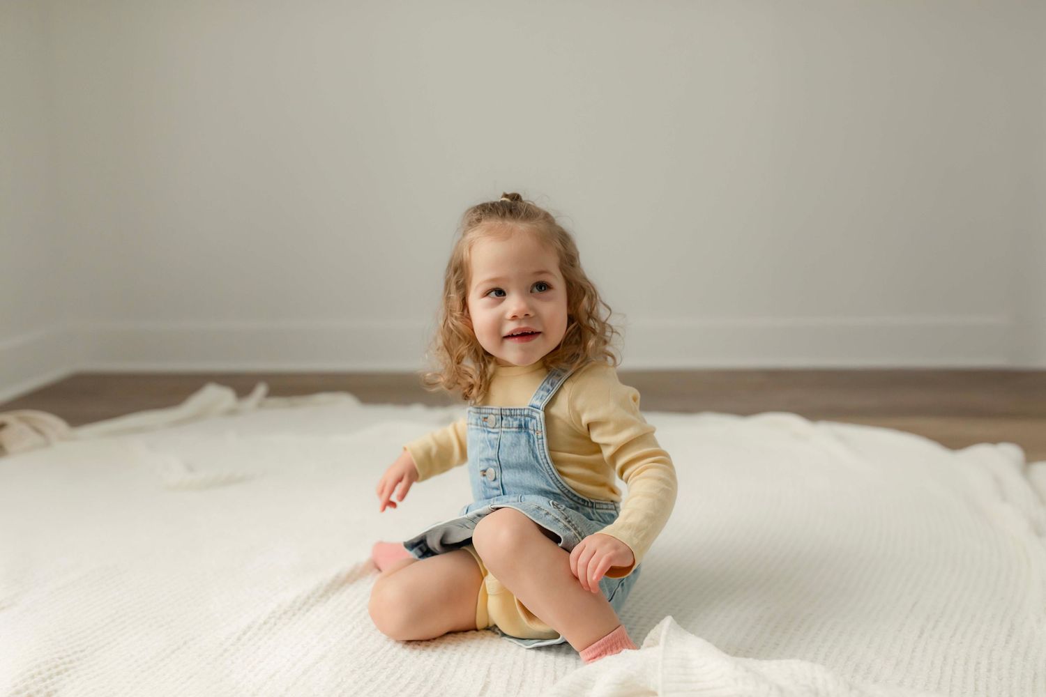 A toddler in beige clothing plays on a white bed in a minimalist room setting.