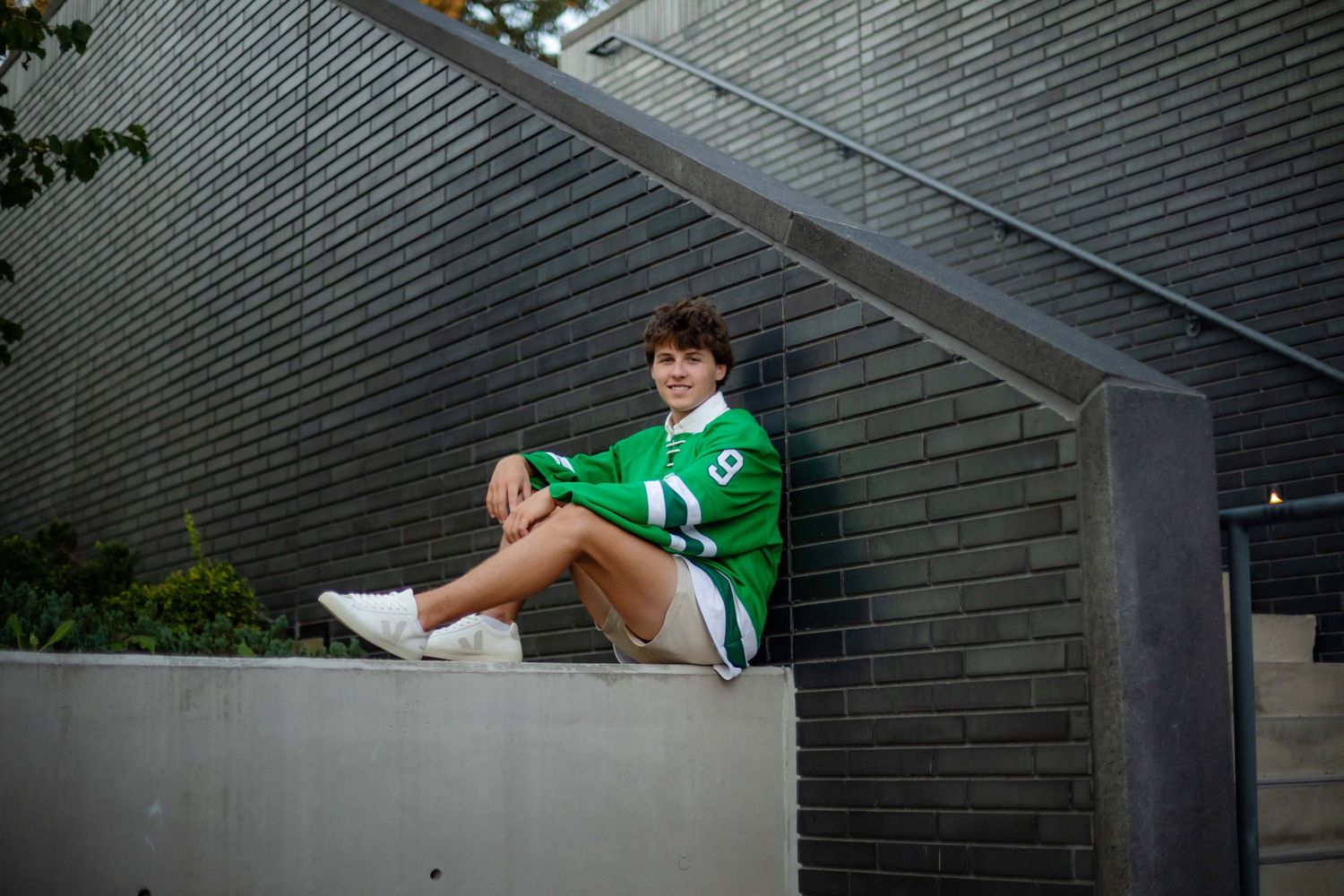 Young man in green jersey sitting on concrete ledge against brick wall outdoors.
