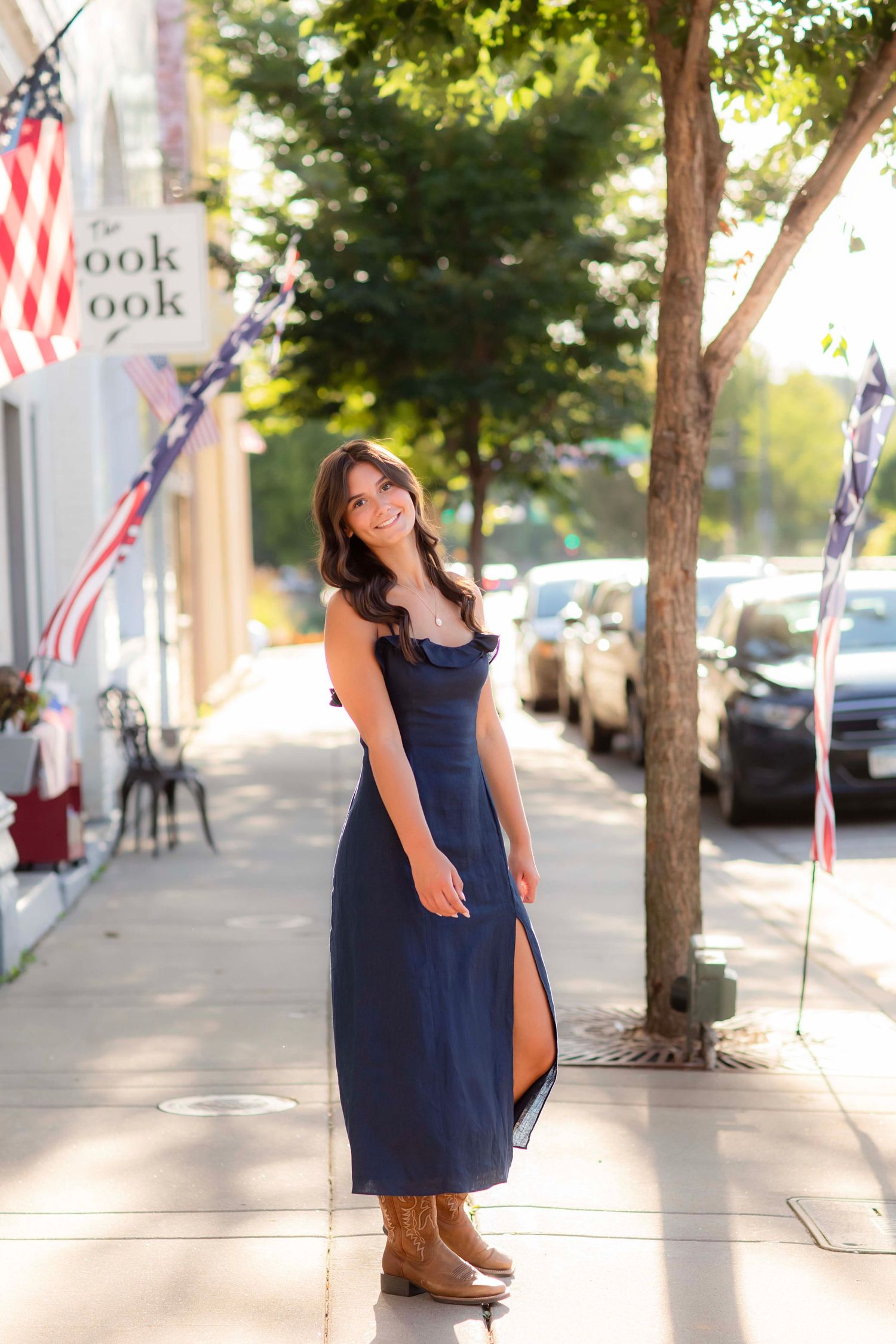Young woman in navy maxi dress and cowboy boots smiling on a sunny small-town sidewalk with American flags.