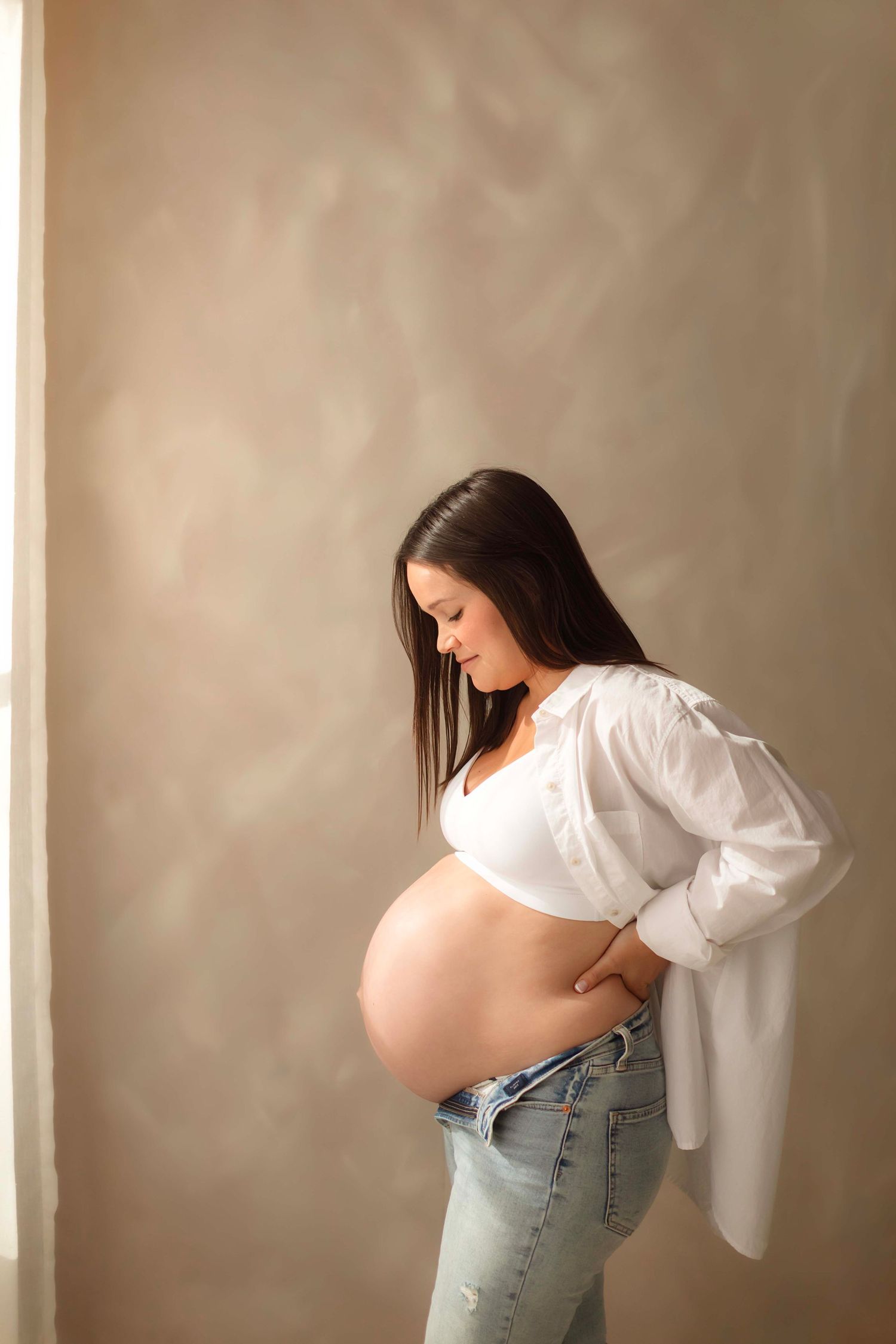 Pregnant woman in white shirt and jeans cradling bare baby bump, looking down lovingly in a softly lit studio.