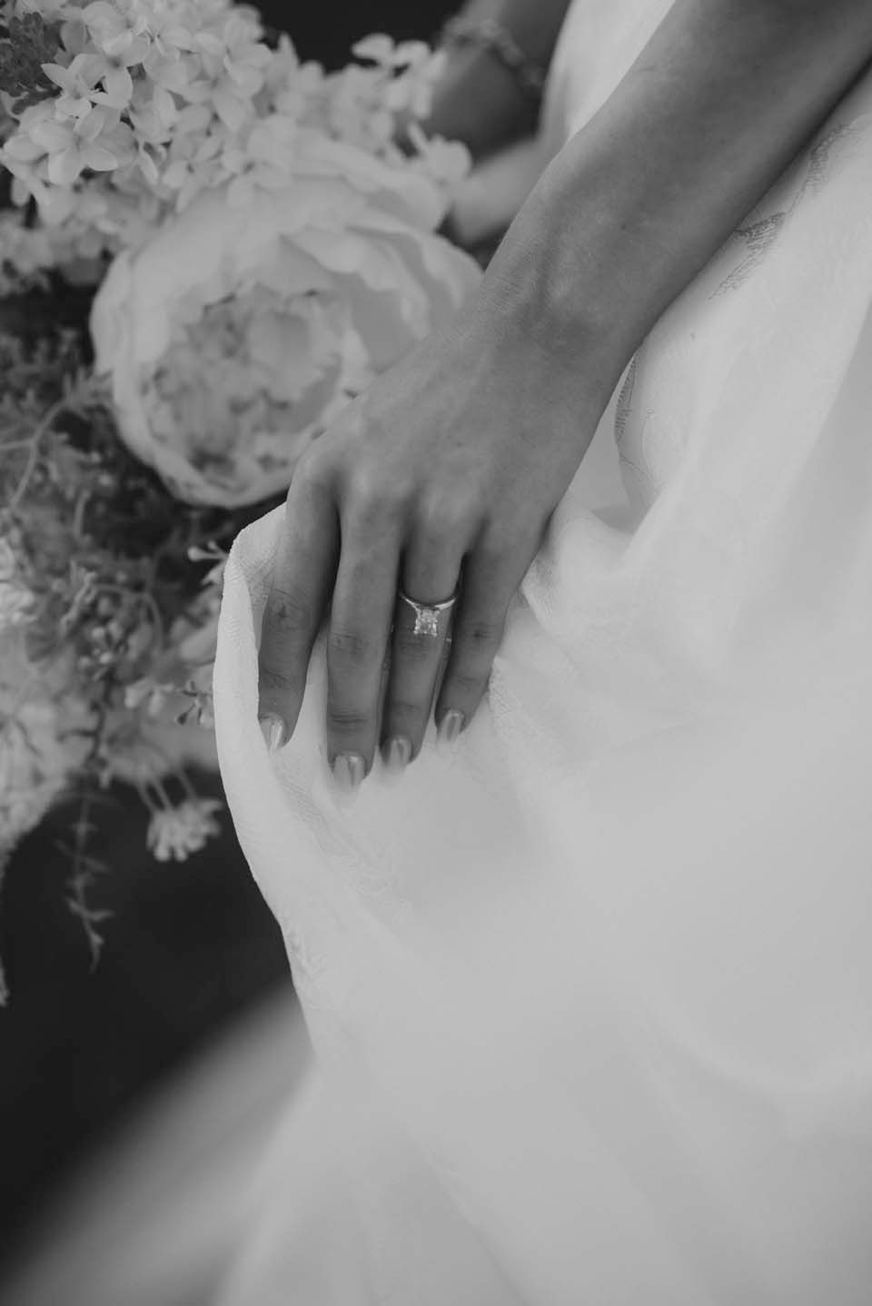 Close-up black and white photo of a bride's hand with an elegant engagement ring resting on her white wedding gown.