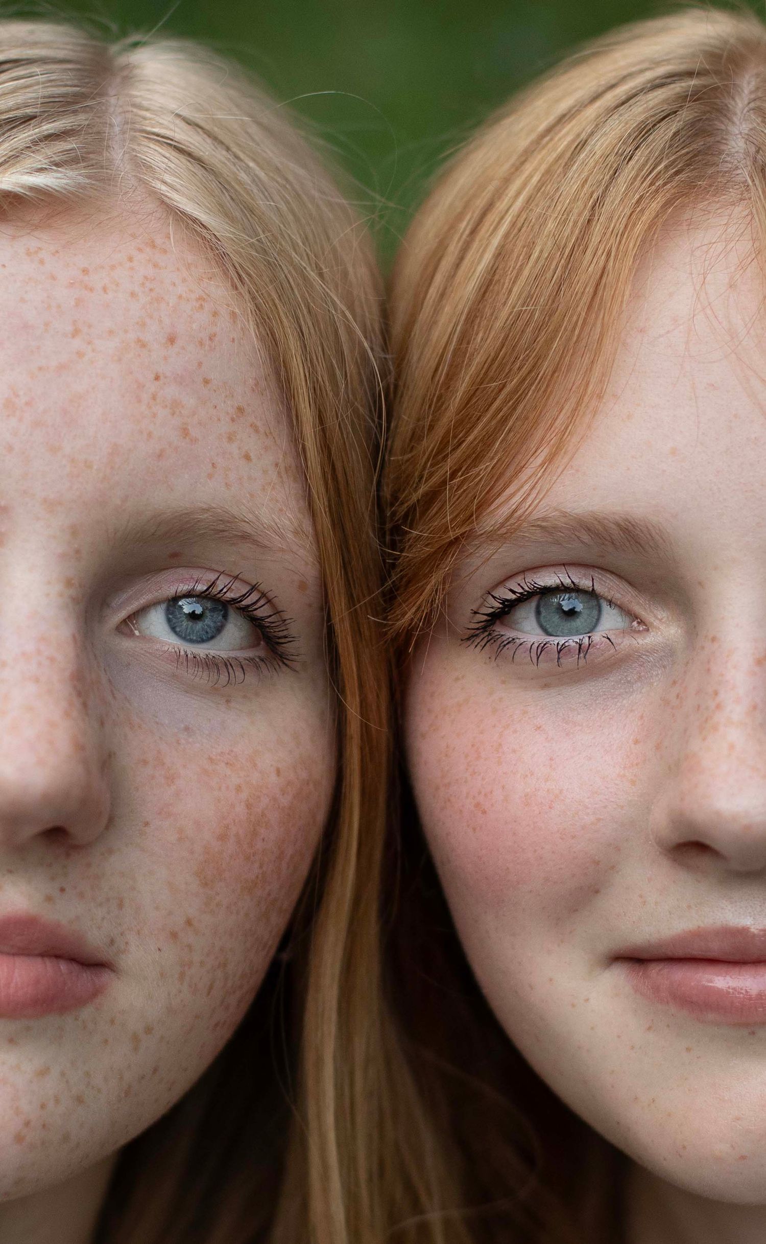 Two young red-haired freckled girls with blue eyes pose cheek to cheek, likely twins, against a green background.