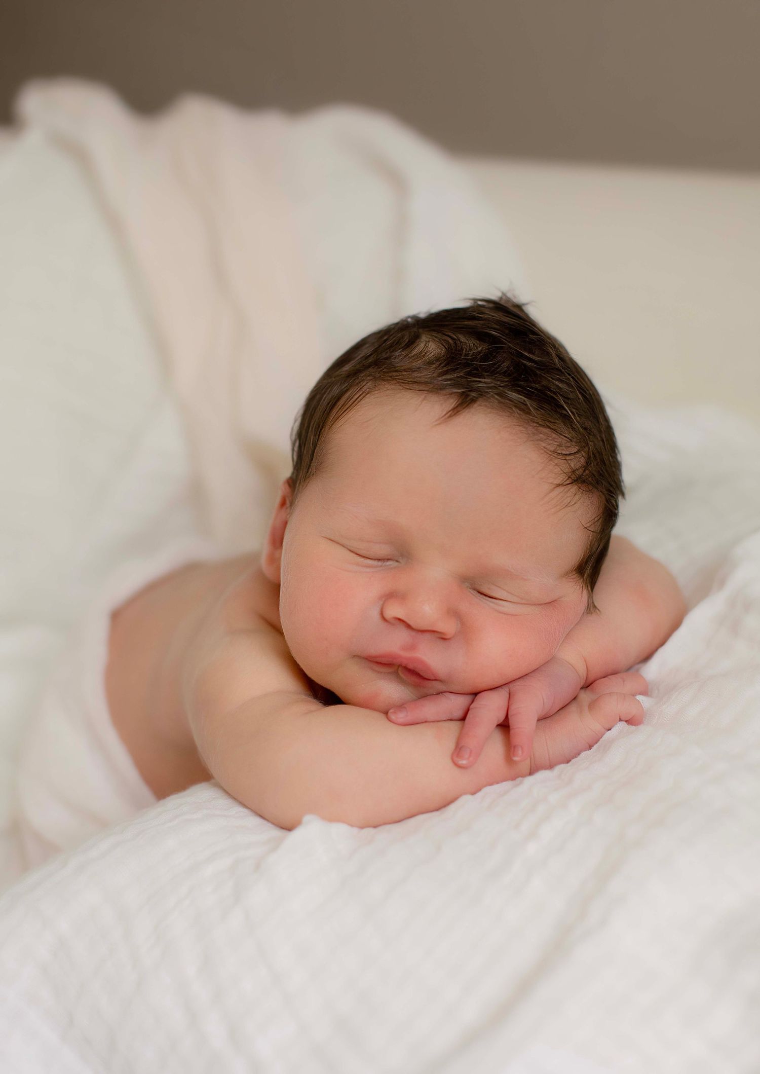 Peaceful sleeping newborn baby with dark hair resting chin on hands on white blanket.