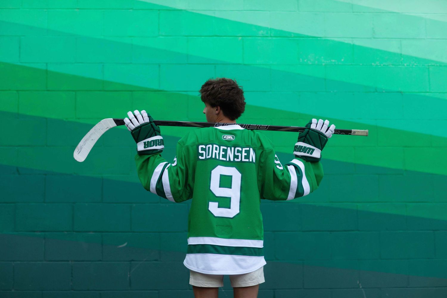 Hockey player wearing green Sorensen #9 jersey holds stick across shoulders, standing against green wall.